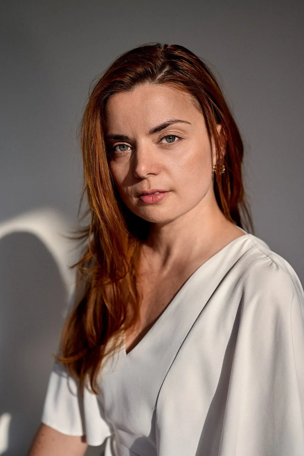 Portrait of a woman with red hair and green eyes wearing a white top, looking at the camera against a gray background.