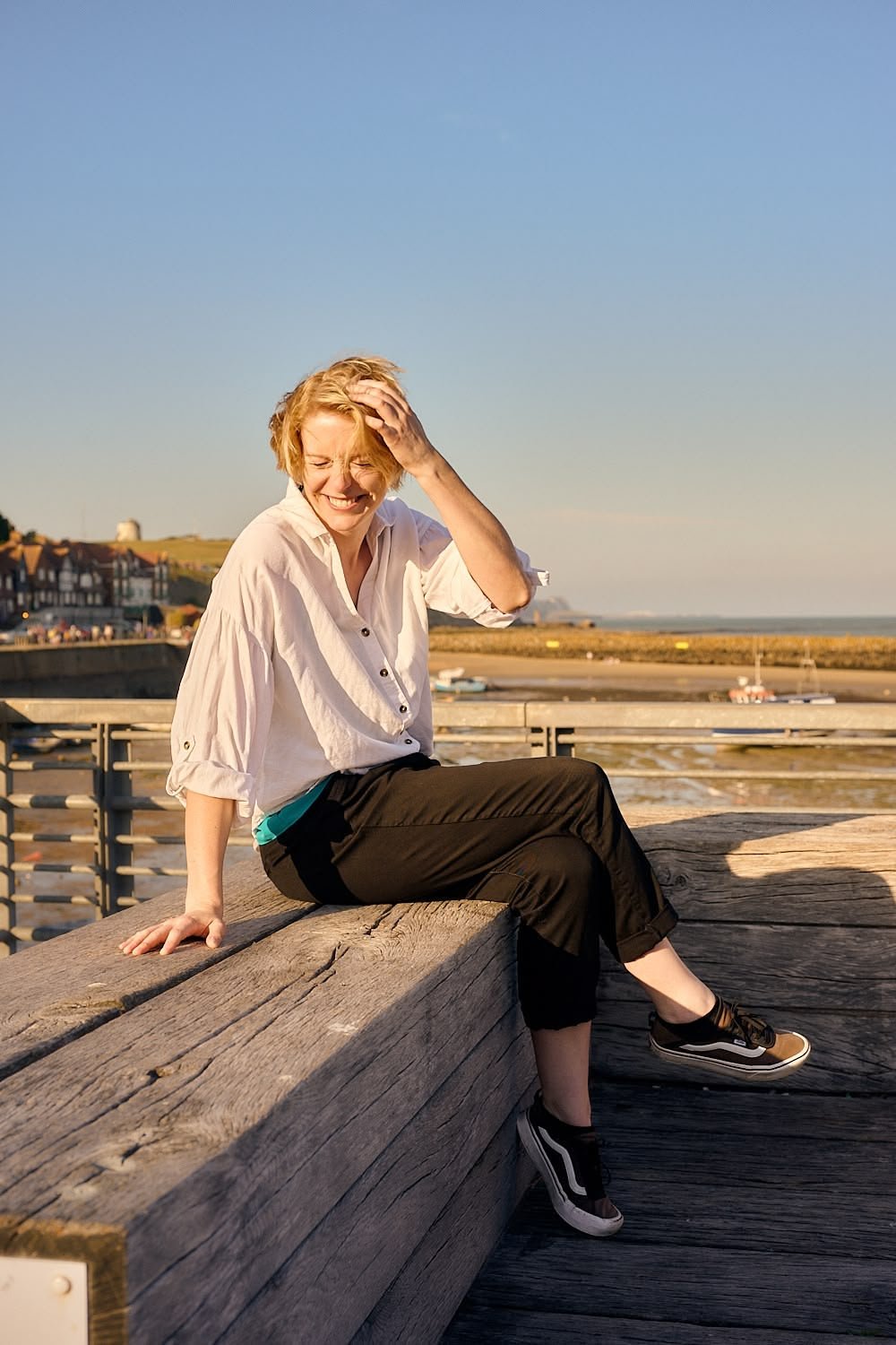 A woman sitting on a wooden bench near a railing at the beach, smiling and laughing while touching her head, with ocean and coastline in the background during sunset.