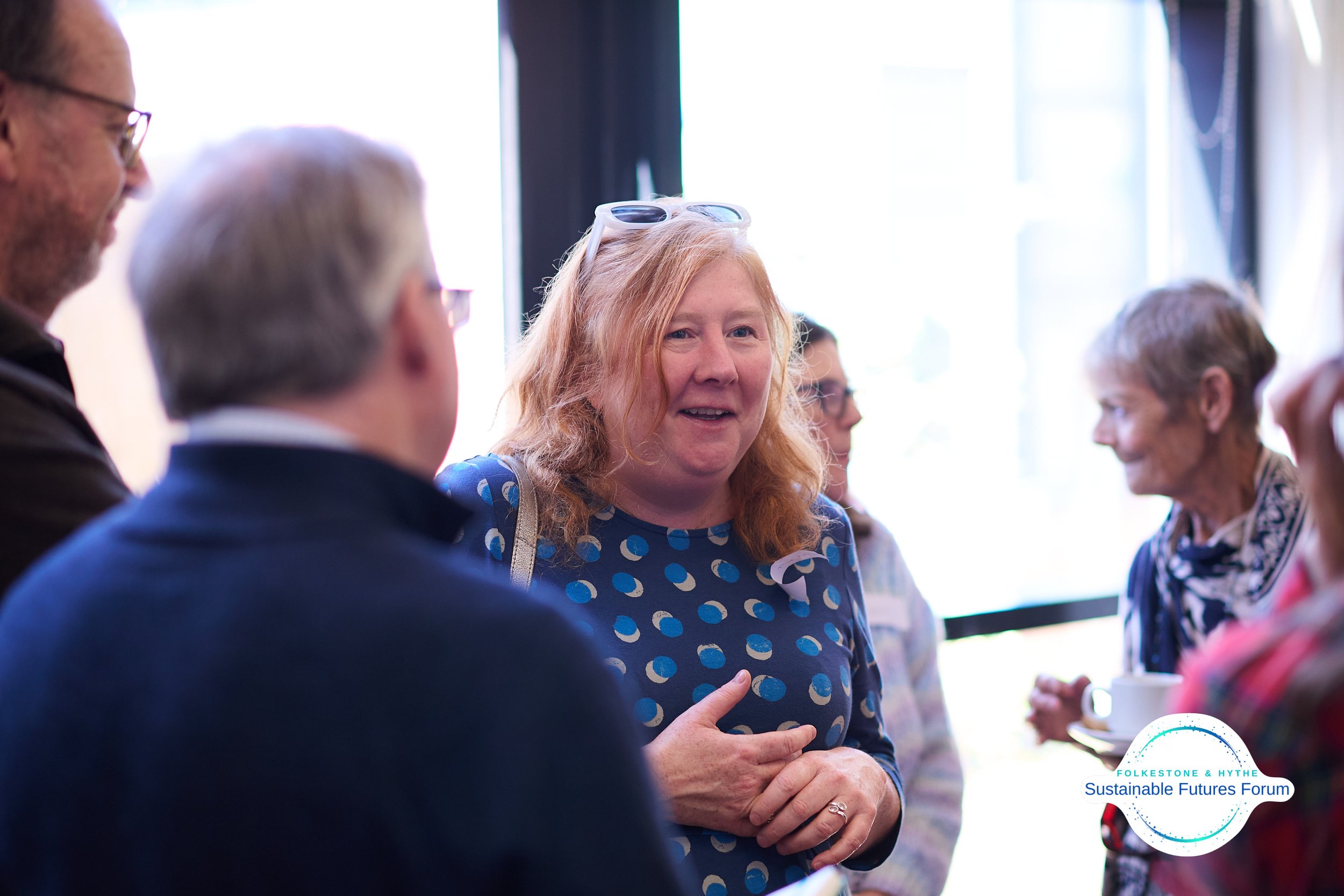 A group of people engaged in conversation at the Sustainable Futures Forum, including a woman with Red hair wearing a blue dress with white and blue polka dots, indoors near a window.