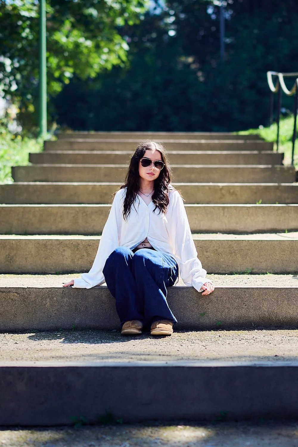 A woman with long dark hair wearing sunglasses, a white long-sleeve shirt, and loose blue jeans sitting on outdoor concrete steps under trees on a sunny day.
