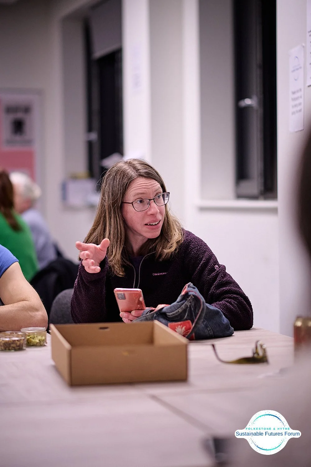 A woman wearing glasses, holding a smartphone, gesturing with her hand while engaging in conversation at a meeting or discussion. There is a cardboard box and some small bowls of snacks on the table. The setting appears to be an indoor community or m