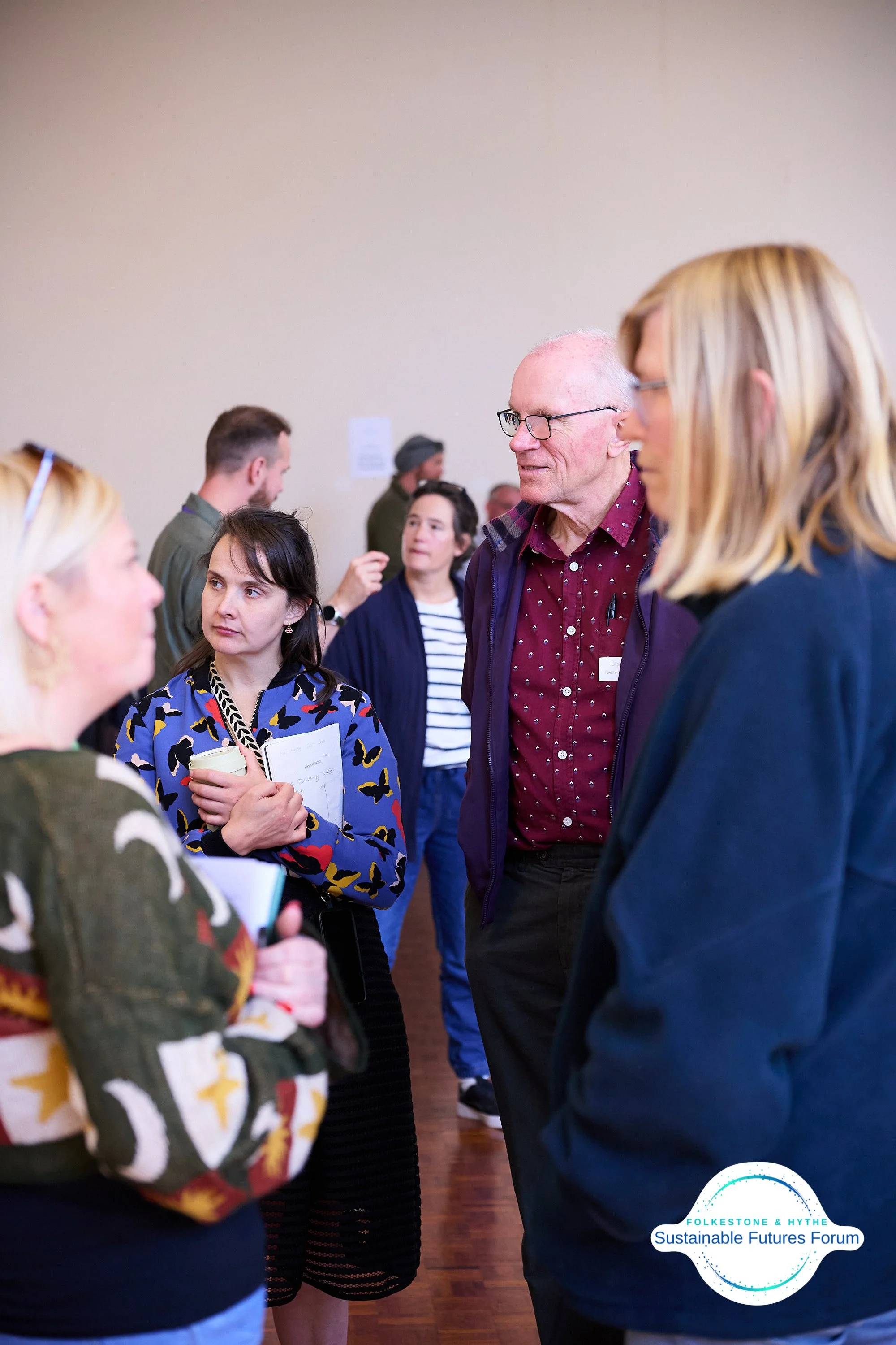 People engaged in conversation at the Folkestone & Hythe Sustainable Futures Forum, with a group of four individuals prominently in focus, conversing indoors.