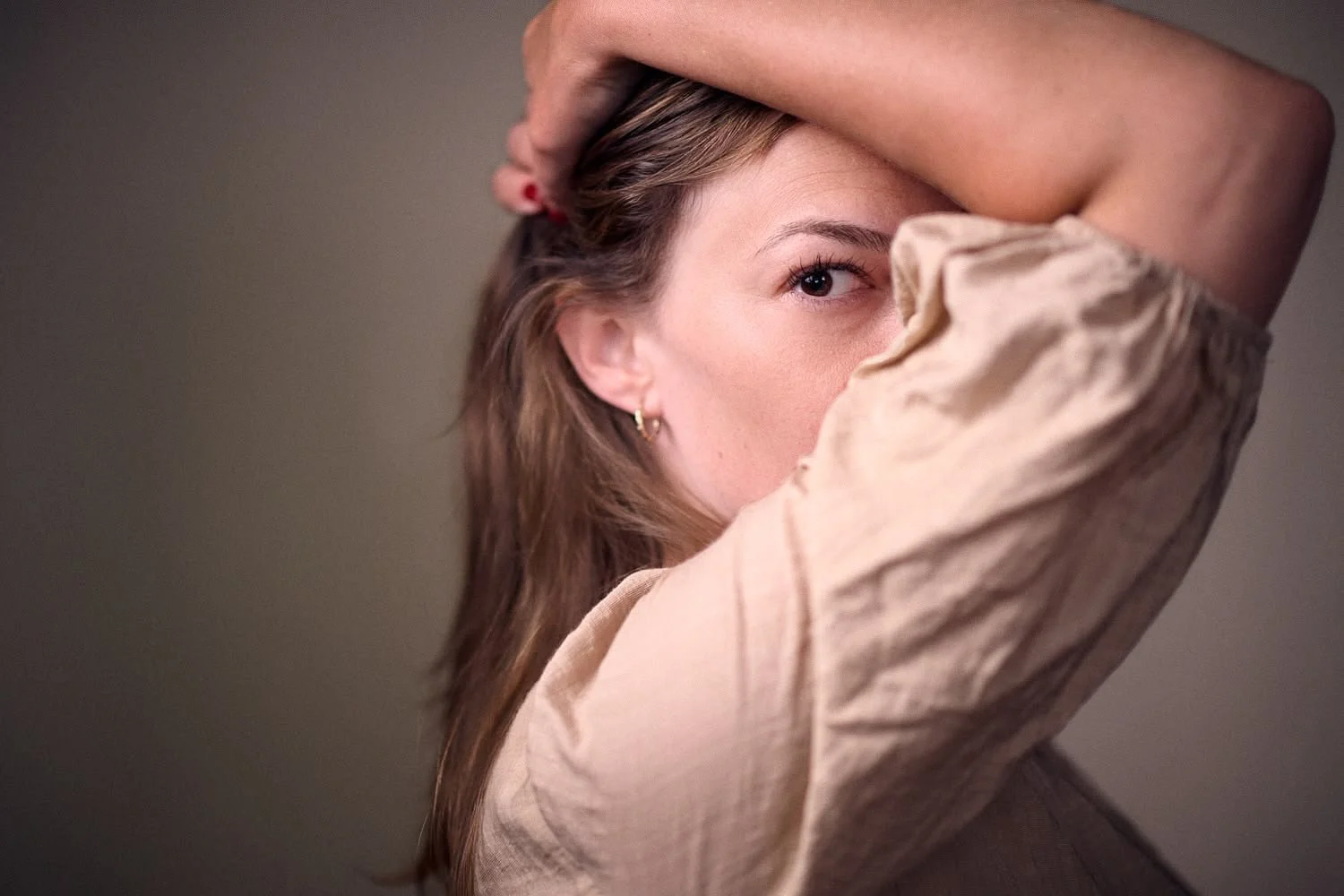 A woman with brown hair posing with one arm over her head, wearing a beige top and gold earrings, looking at the camera with one eye visible against a plain background.