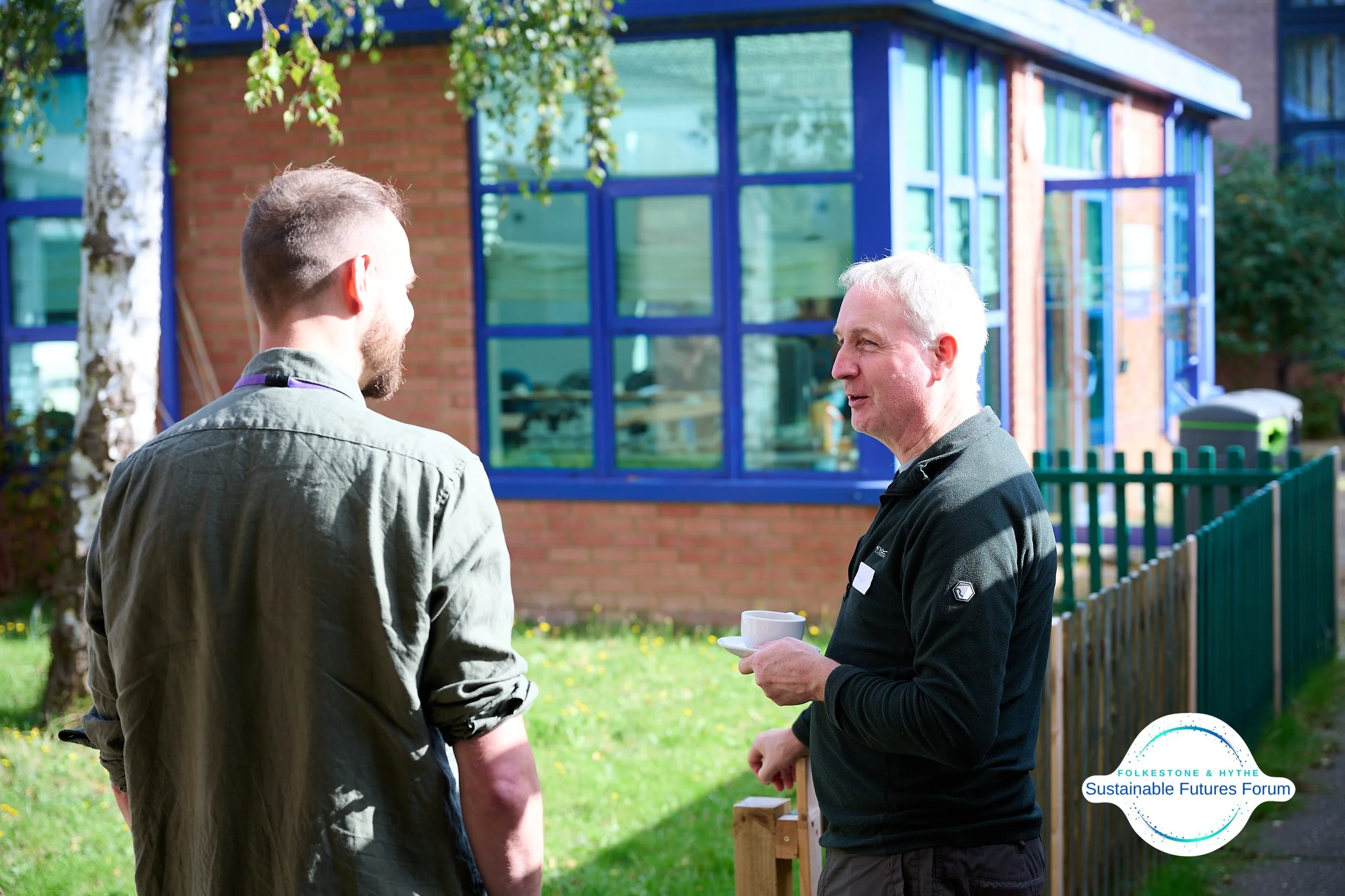 Two men having a conversation outdoors on a sunny day, with a building featuring large blue-framed windows in the background, and the logo of FolkeStone & Hythe Sustainable Futures Forum in the bottom right corner.