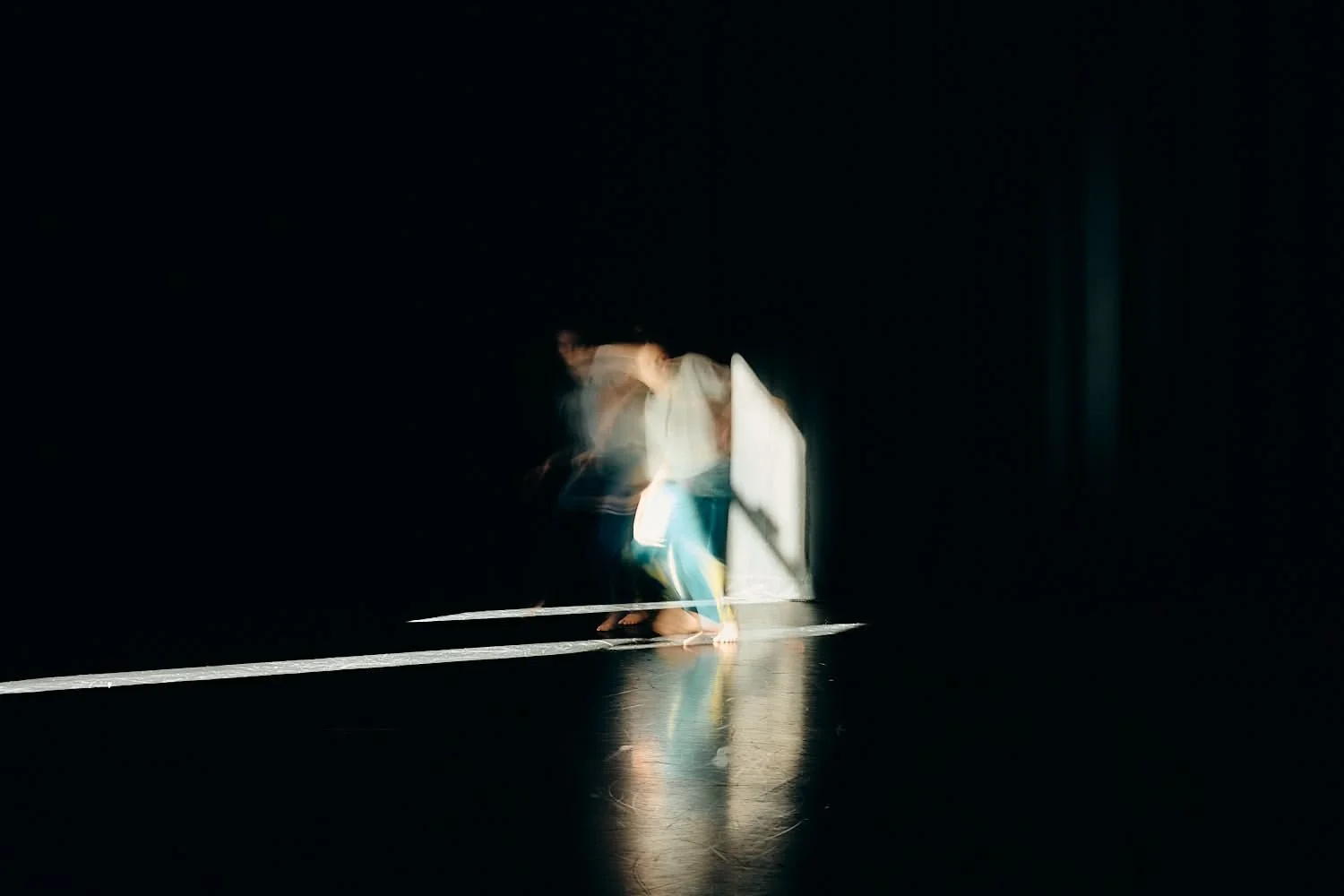 A blurred image of a person walking on a dark stage or floor, with bright light illuminating the person's lower body and feet, creating reflections on the floor.