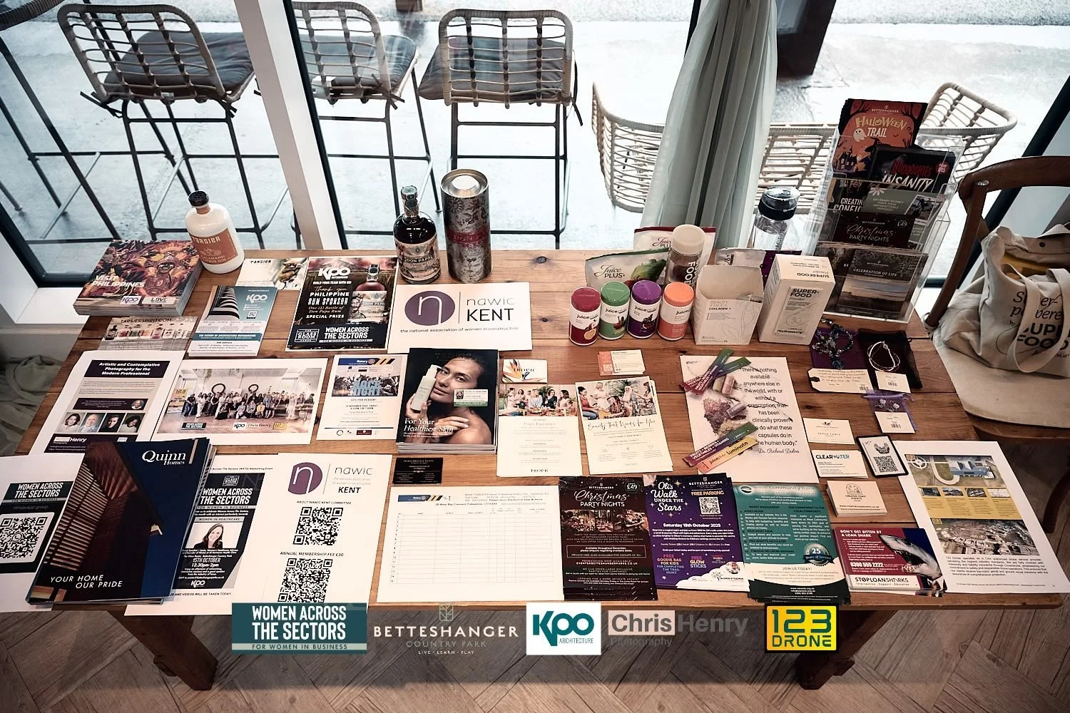 A wooden table displaying various flyers, brochures, promotional materials, and some small bottles, with a large window in the background showing outdoor chairs and a patio.