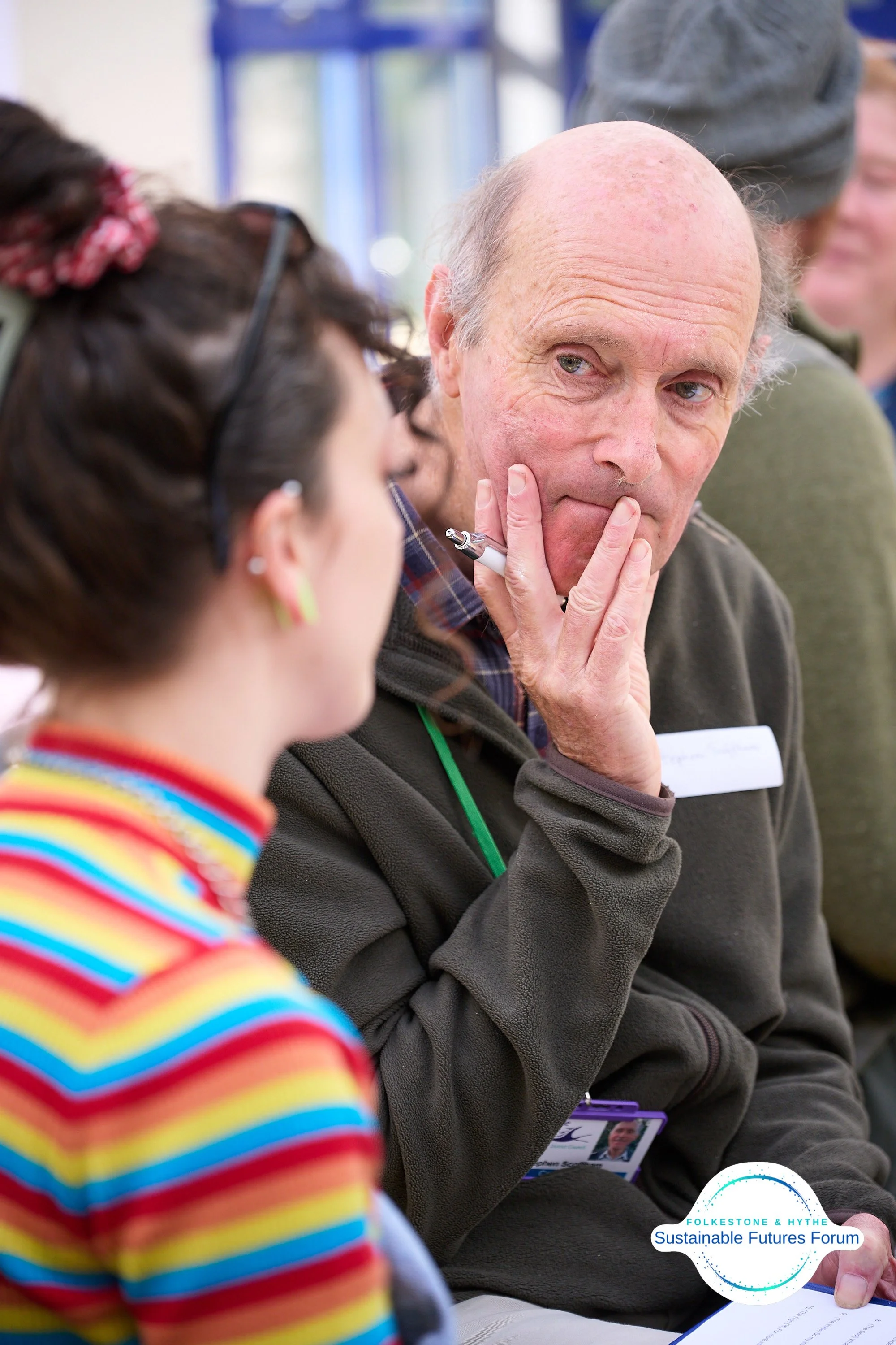 An older man with a bald head and light skin holding a pen to his face, engaged in conversation with a woman with dark hair and colorful striped clothing at a forum, with a logo for Folkesone & Hythe Sustainable Futures Forum in the bottom right corn