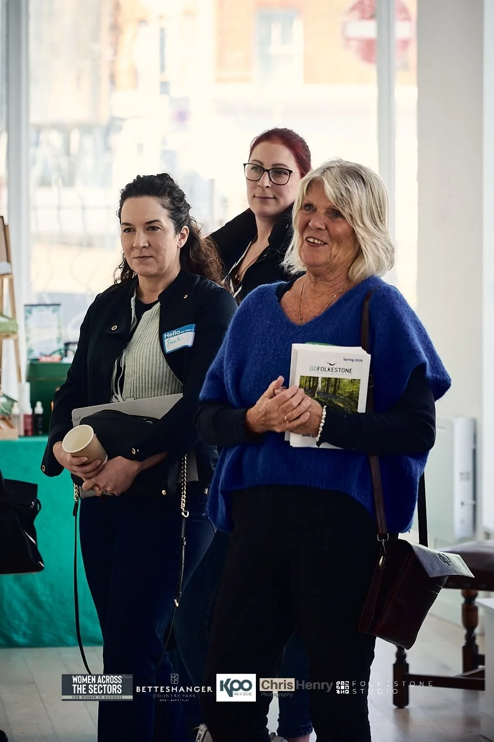 Three women standing indoors during a daytime event. The woman in the foreground wears a blue sweater and holds a pamphlet, smiling. The woman behind her has blonde hair, glasses, and wears a black jacket. The third woman, with dark curly hair, holds
