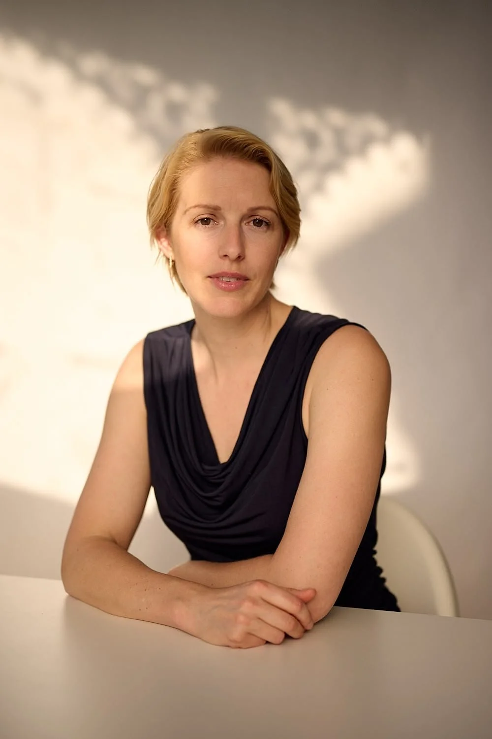 A woman with short blonde hair sitting at a table with a neutral expression, wearing a sleeveless black top, with a plain background.