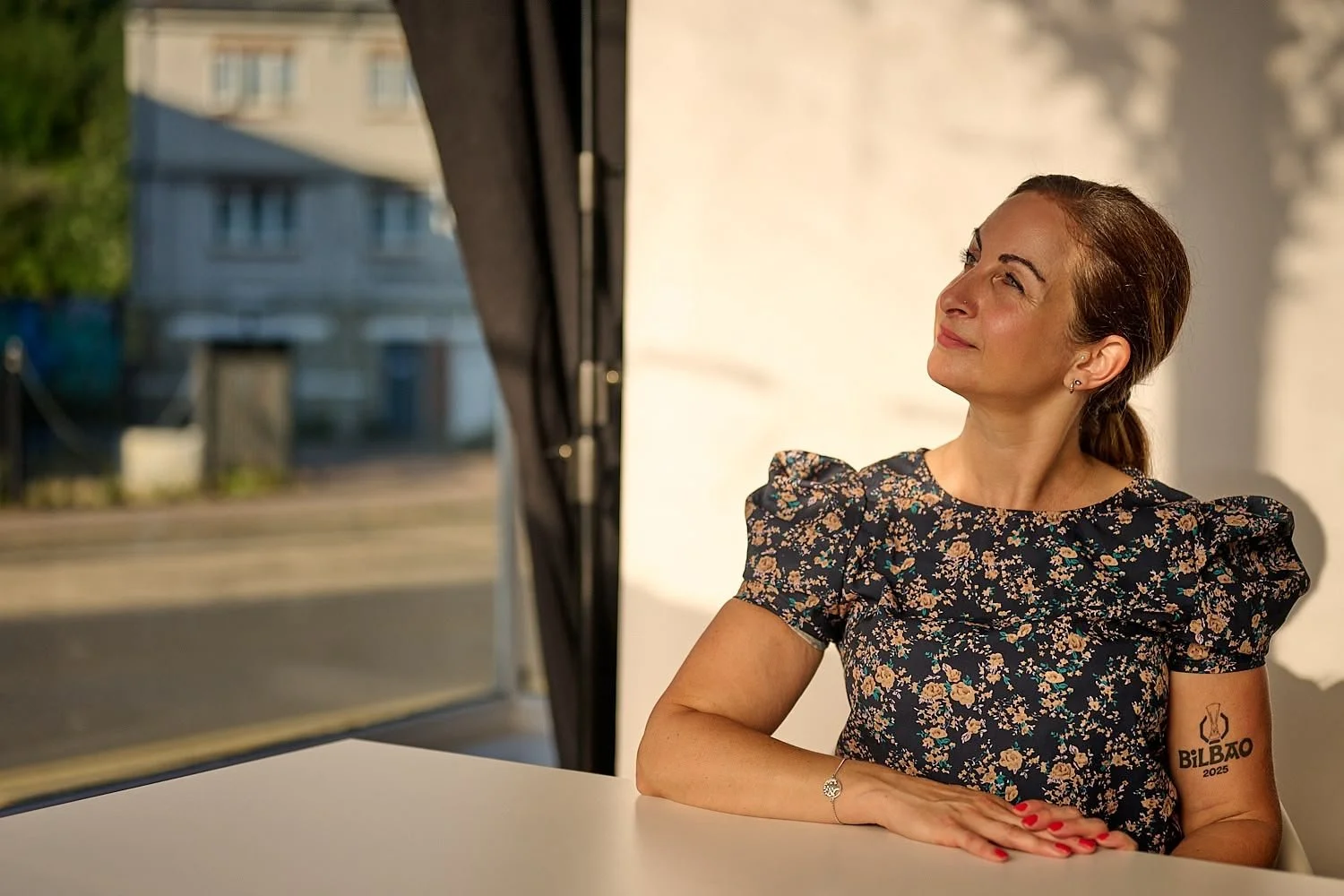 A woman with brown hair pulled back sitting at a white table, looking outside through a window on a sunny day, wearing a dark floral dress and a bracelet on her left wrist.