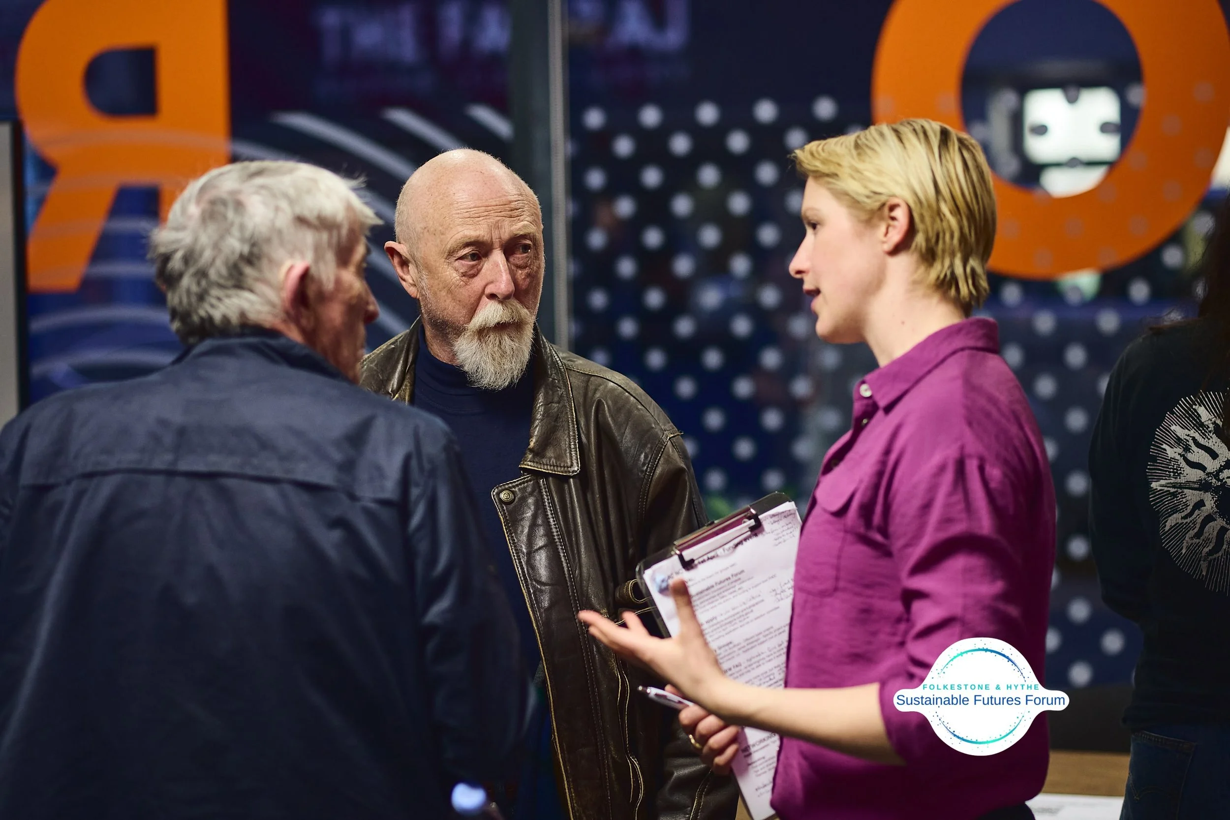 Three people speaking at a forum, one woman holding a clipboard, others listening, with a 'Sustainable Futures Forum' logo in the bottom right corner.