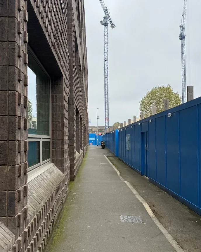 Sidewalk next to a brick building and a blue construction fence, with cranes visible in the background on a cloudy day.