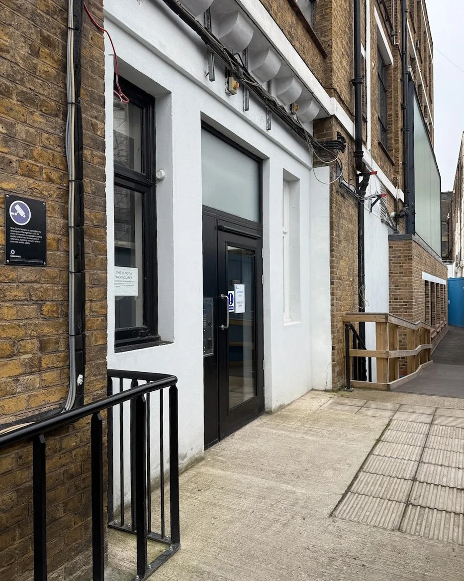 Outside view of a building entrance with a glass door, a black metal handrail, a white wall, and brick structure around the entrance, with signage and electrical conduits on the wall.