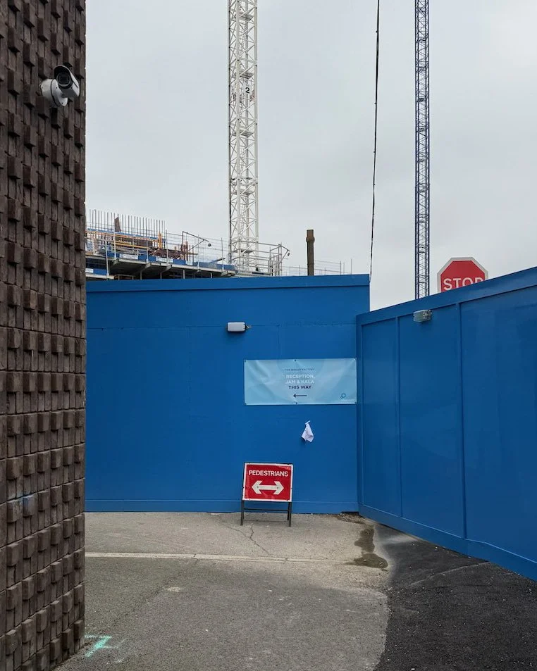 A blue construction site fence with signs indicating pedestrian directions, a stop sign visible in the background, and construction cranes behind the fence.