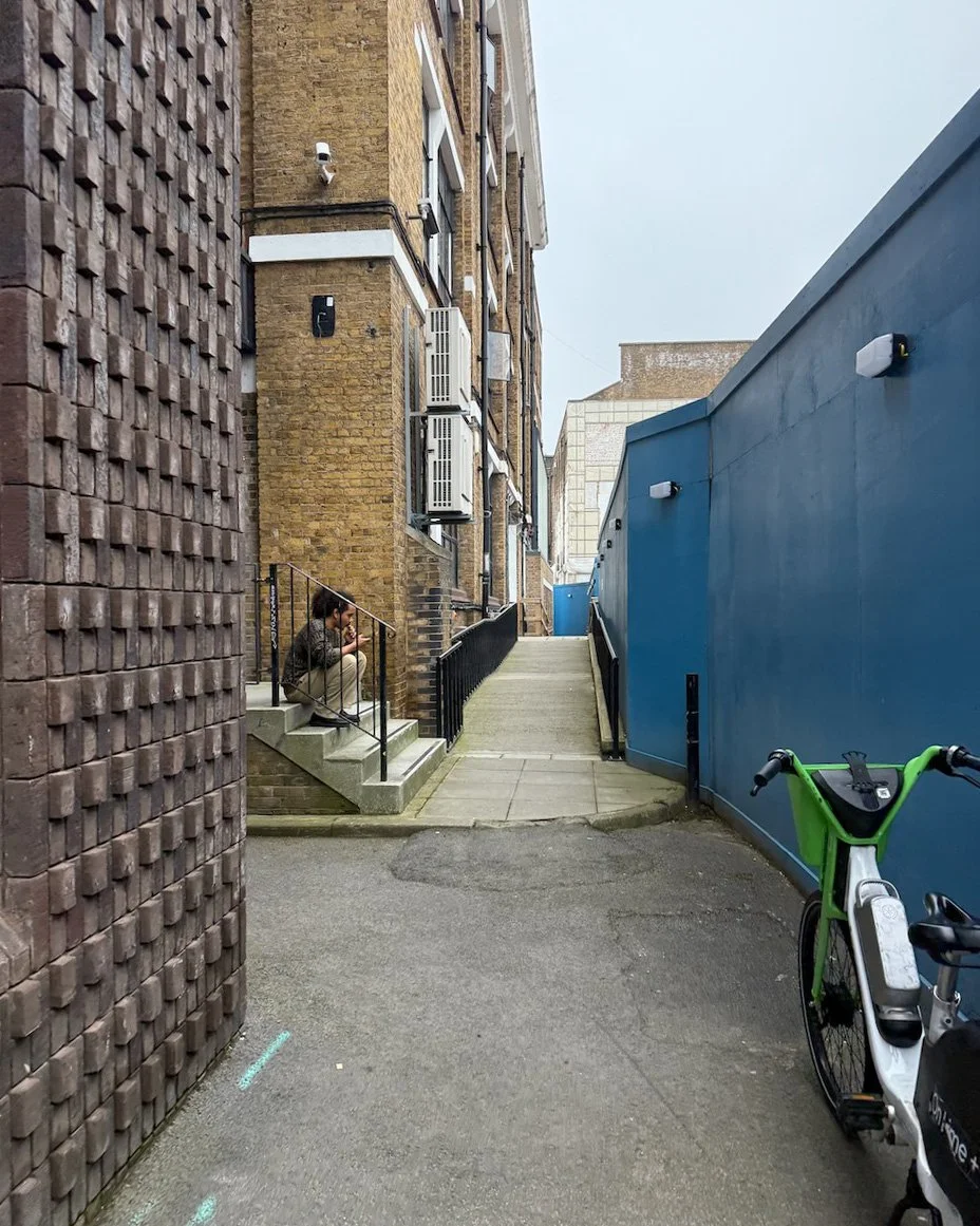 A person sitting on stairs outside a brick building, looking at their phone. There is a blue wall to the right with a green bike parked against it, and a ramp leading up towards more buildings in the background.