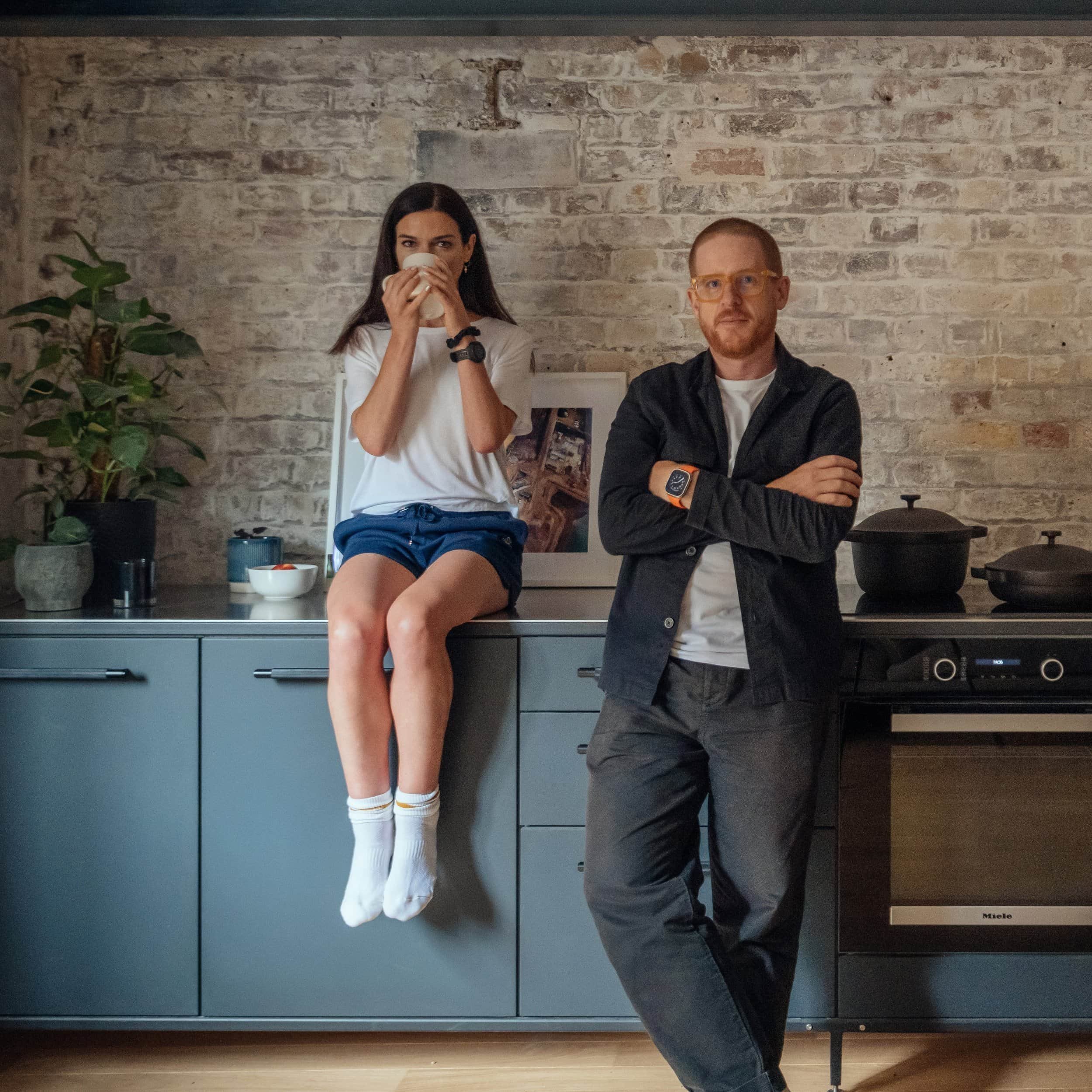 A woman sitting on a kitchen counter drinking from a mug, and a man standing with arms crossed next to her, in a modern kitchen with a brick wall background.