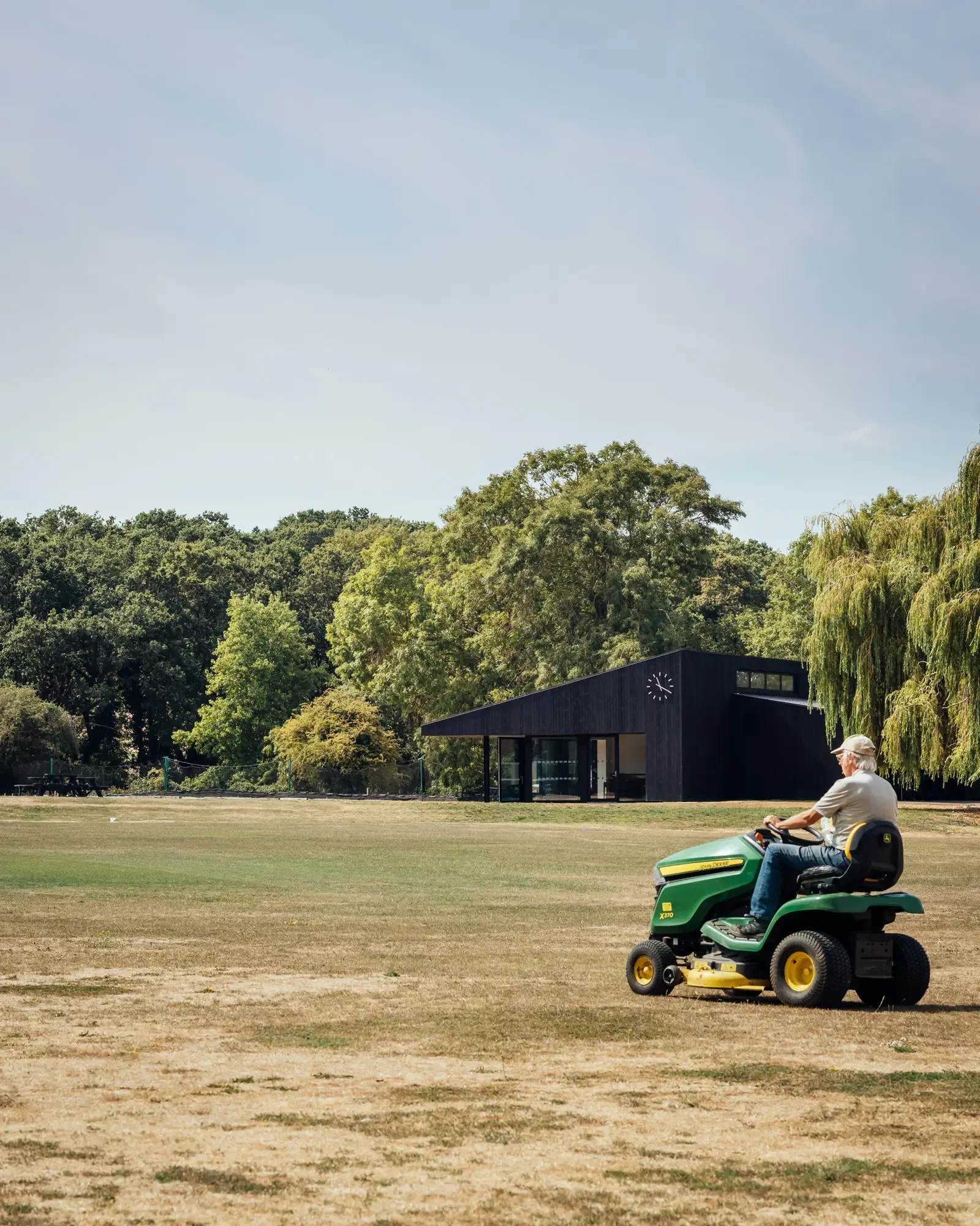 Groundskeeper on ride on mower in foreground with modern cricket pavilion in the background © Jim Stephenson