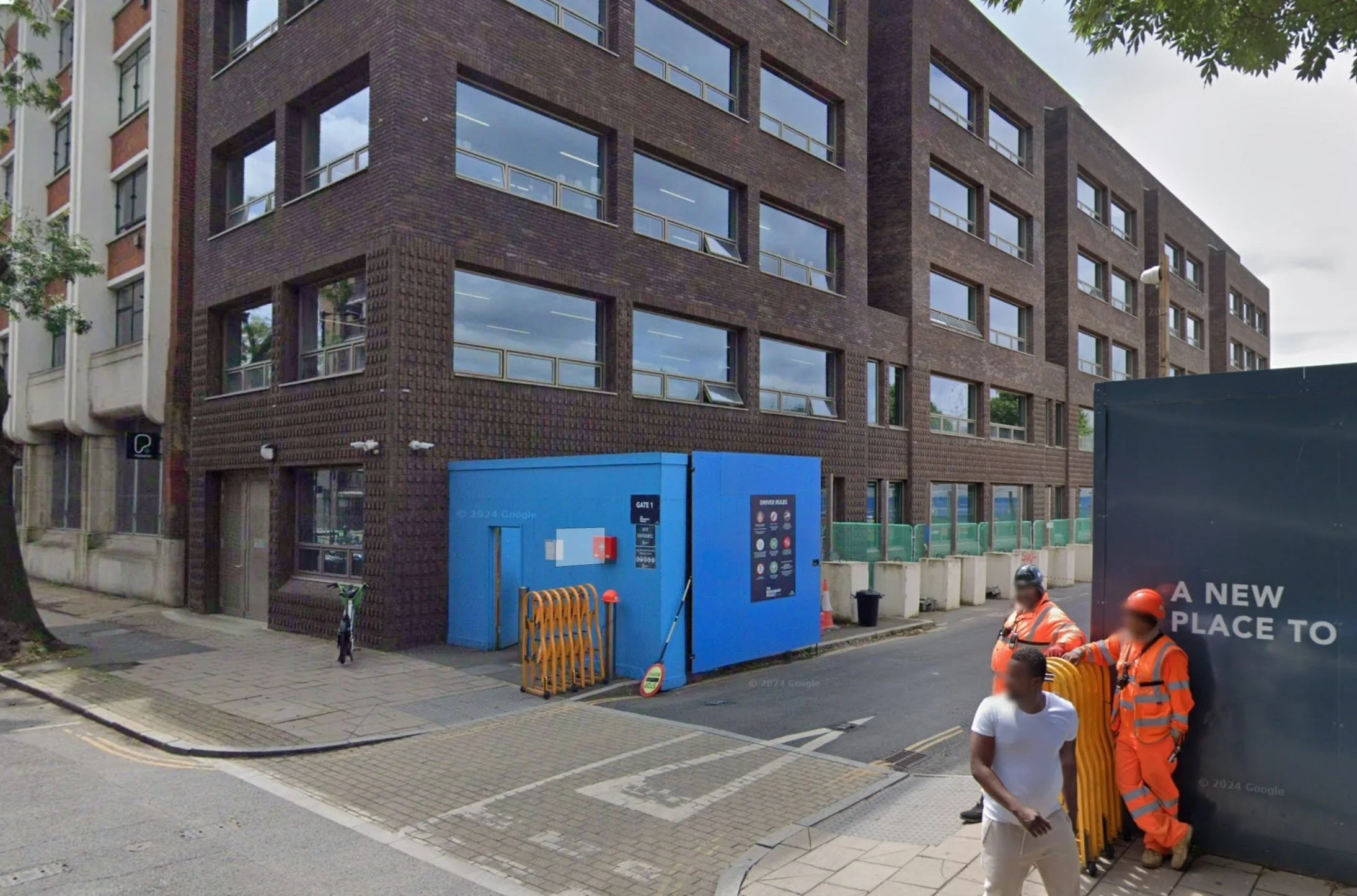 Urban street scene with a multi-story brown brick building, blue construction gate, and construction workers in orange safety vests and helmets.