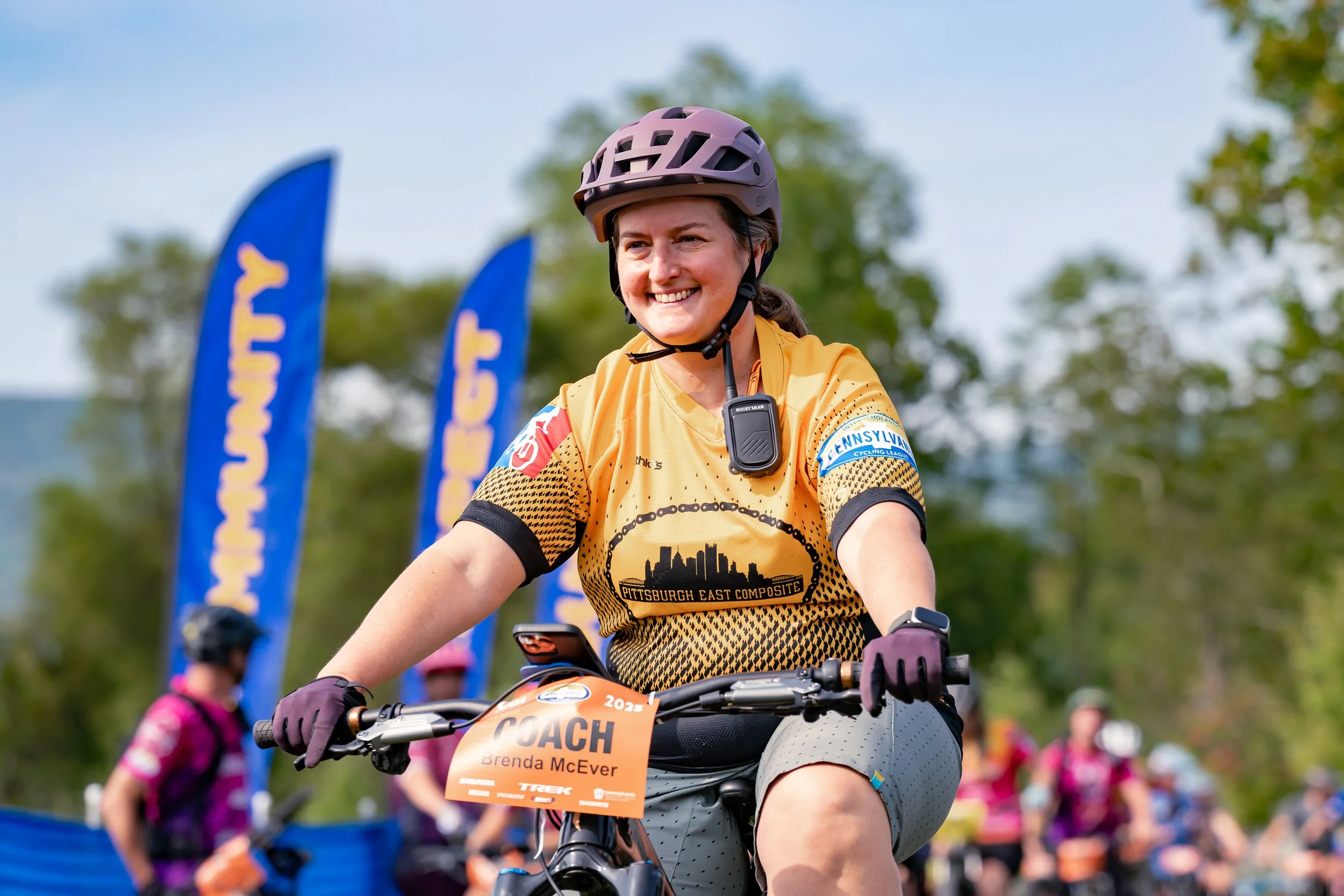 A woman wearing a yellow cycling jersey, helmet, and gloves rides a bike during an outdoor event with blue flags in the background. She is smiling, and a sign on her bike reads 'COACH Brenda McEver'.