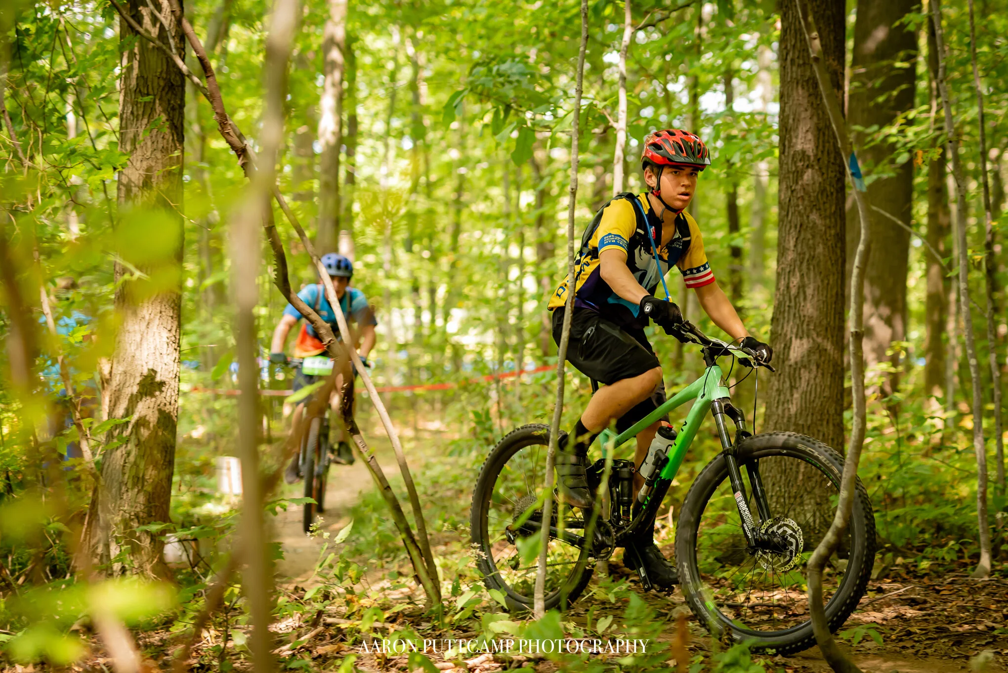 Two young boys riding bicycles through a densely wooded forest, wearing helmets and sports gear.