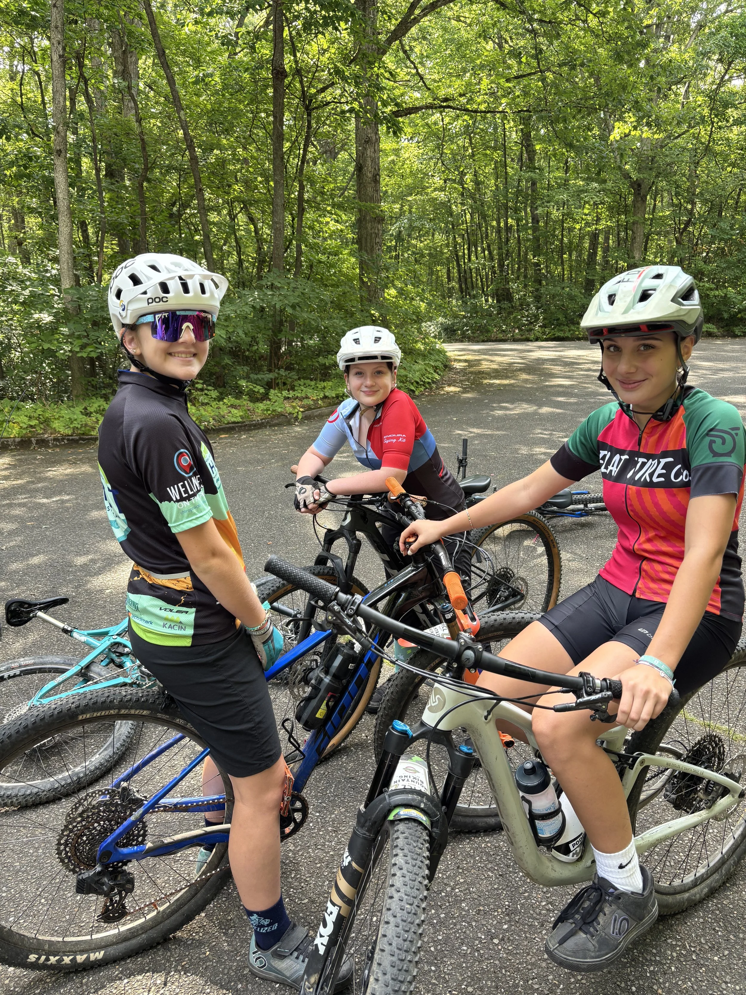Three young girls wearing mountain biking gear and helmets, standing with their bikes on a paved trail surrounded by lush green forest.