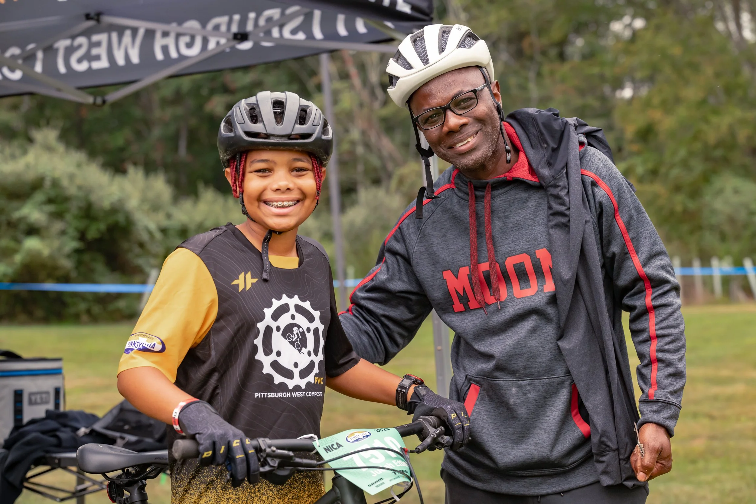 A smiling young girl and an adult man wearing helmets, standing together outdoors with a bicycle. The girl is dressed in a black and yellow cycling jersey, and the man in a gray and red hoodie. They appear to be at a cycling event.