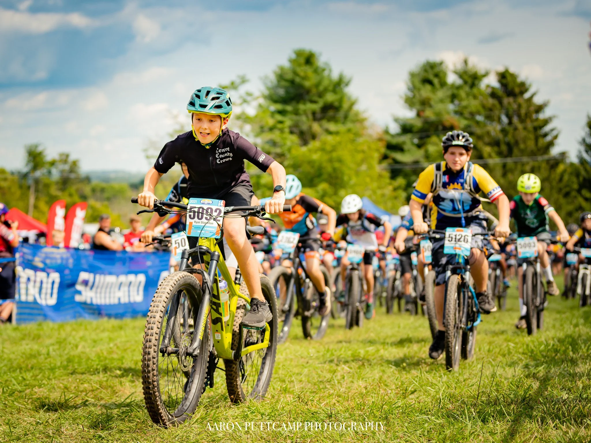 Children participating in a mountain bike race on a grassy field, wearing helmets and racing gear, with a crowd and race banners in the background.