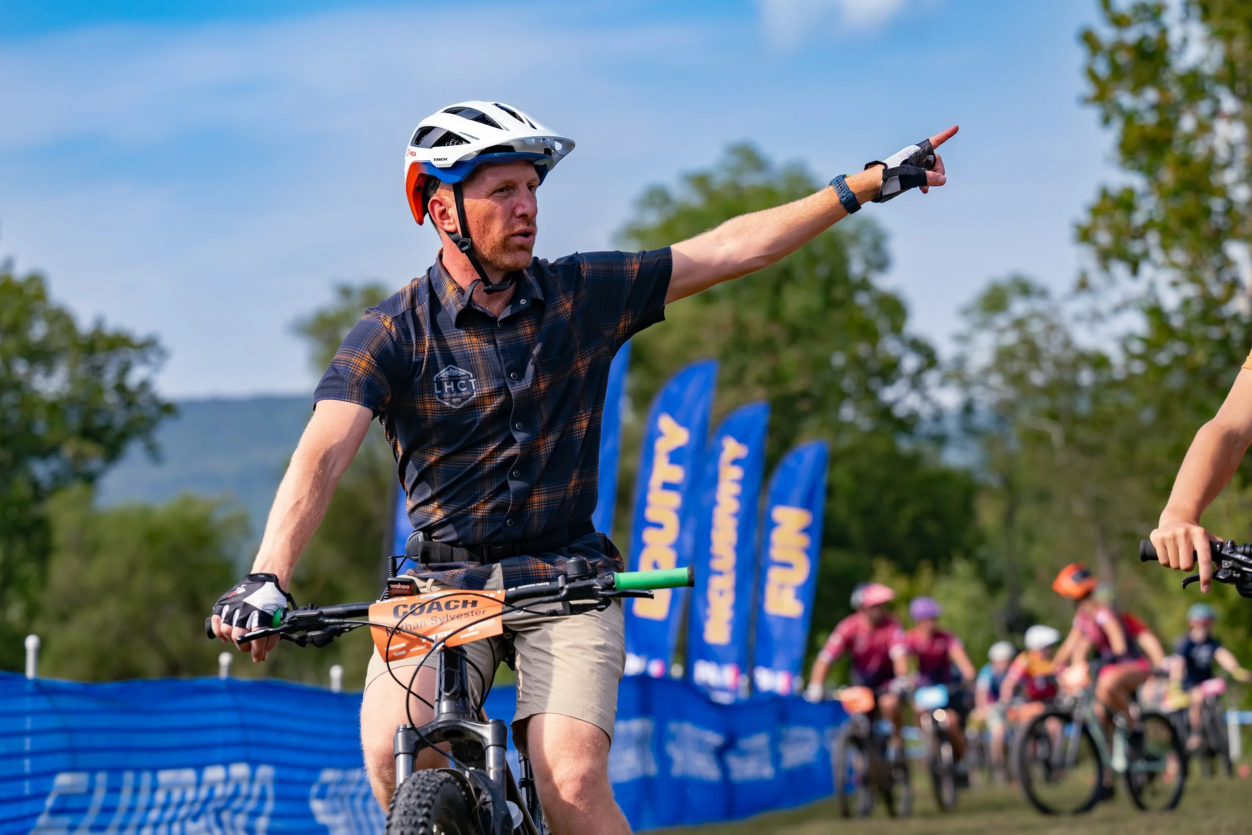 A man wearing a helmet, gloves, and a plaid shirt on a mountain bike, pointing forward during a cycling event with other cyclists in the background, and banners that read 'FUN' and 'RACE' in an outdoor setting.