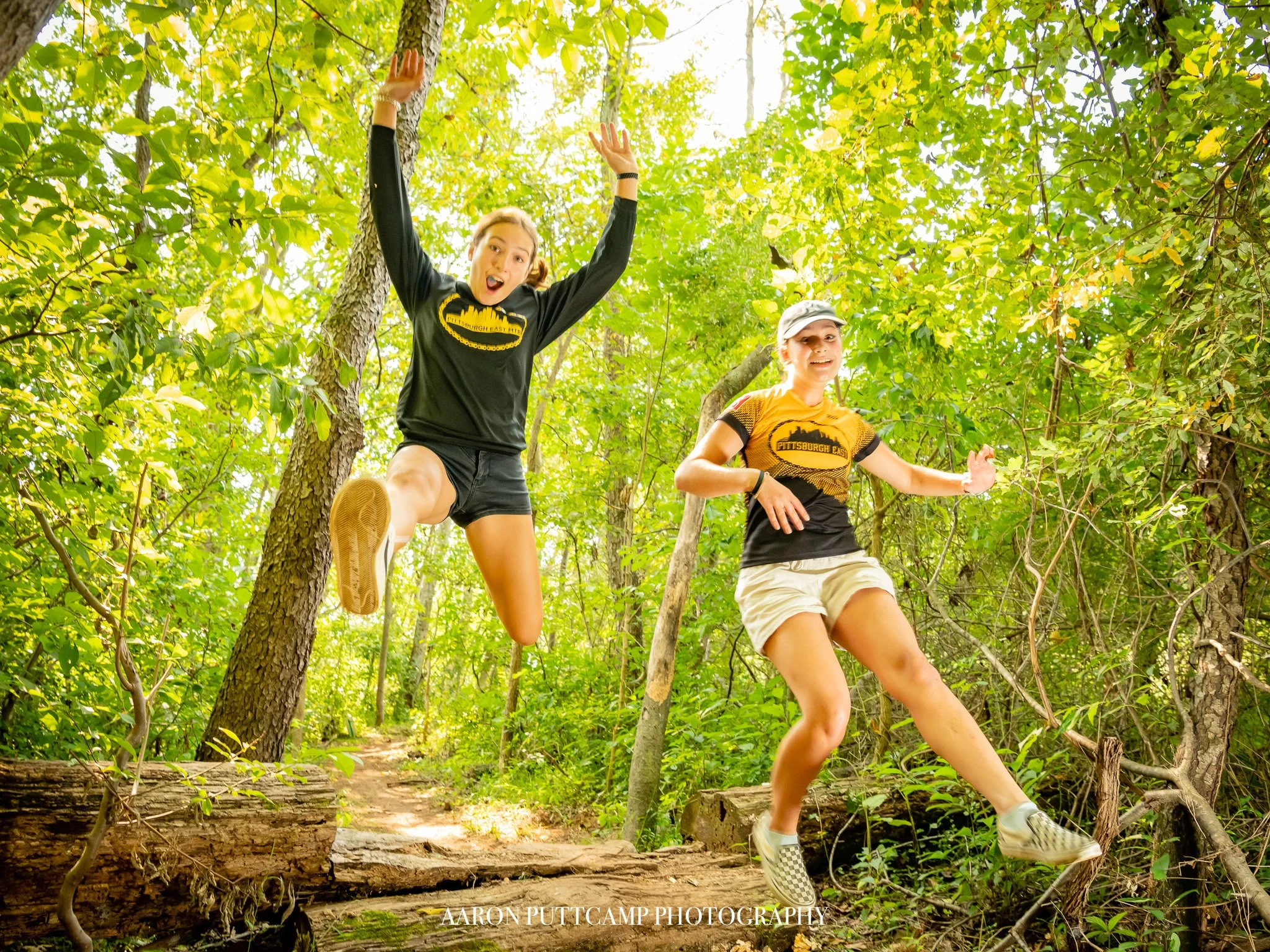 Two women in black, yellow, and gray athletic clothing are jumping and running on a forest trail surrounded by green foliage.