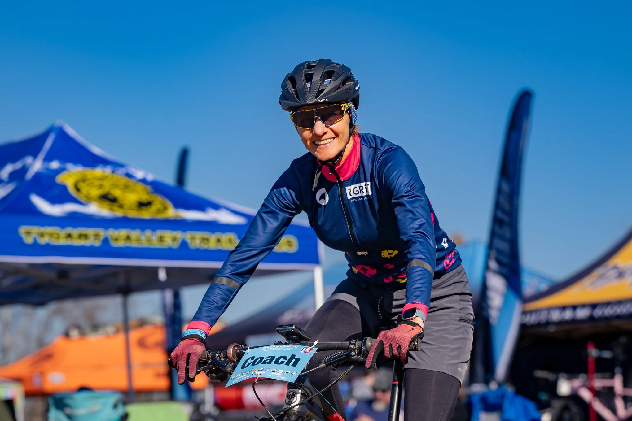A woman wearing a helmet, sunglasses, a blue long-sleeve cycling jersey, and gloves, smiling while riding a bicycle at an outdoor event with tents and flags in the background.