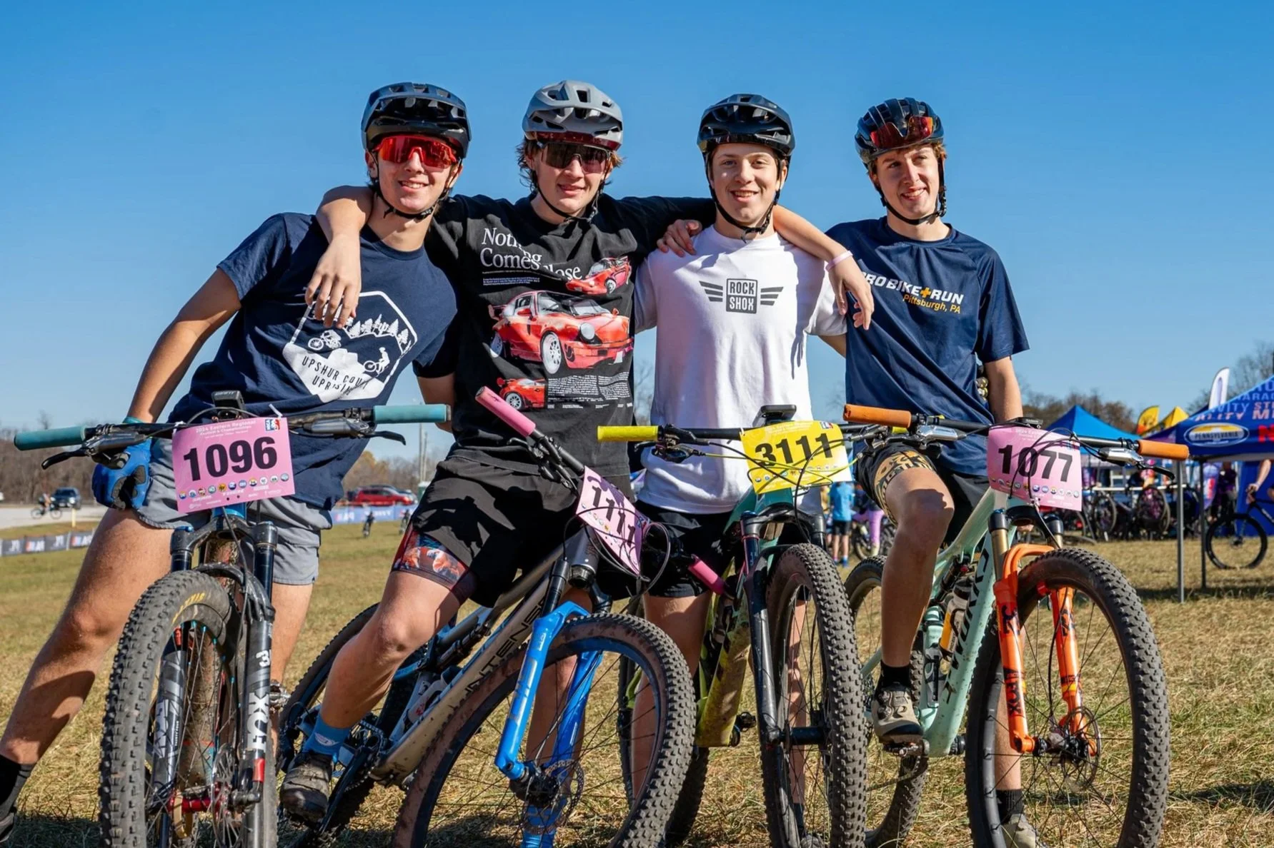 Four young boys with helmets and sunglasses, standing with their mountain bikes on a grassy field during a biking event, smiling and posing for the camera.