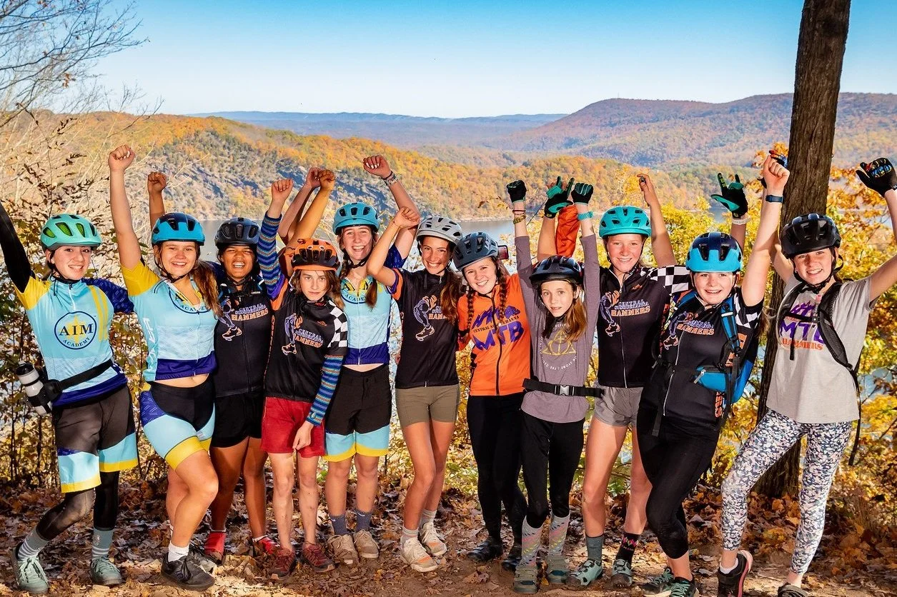 Group of young female cyclists raising their arms in celebration outdoors with fall foliage and mountains in the background.