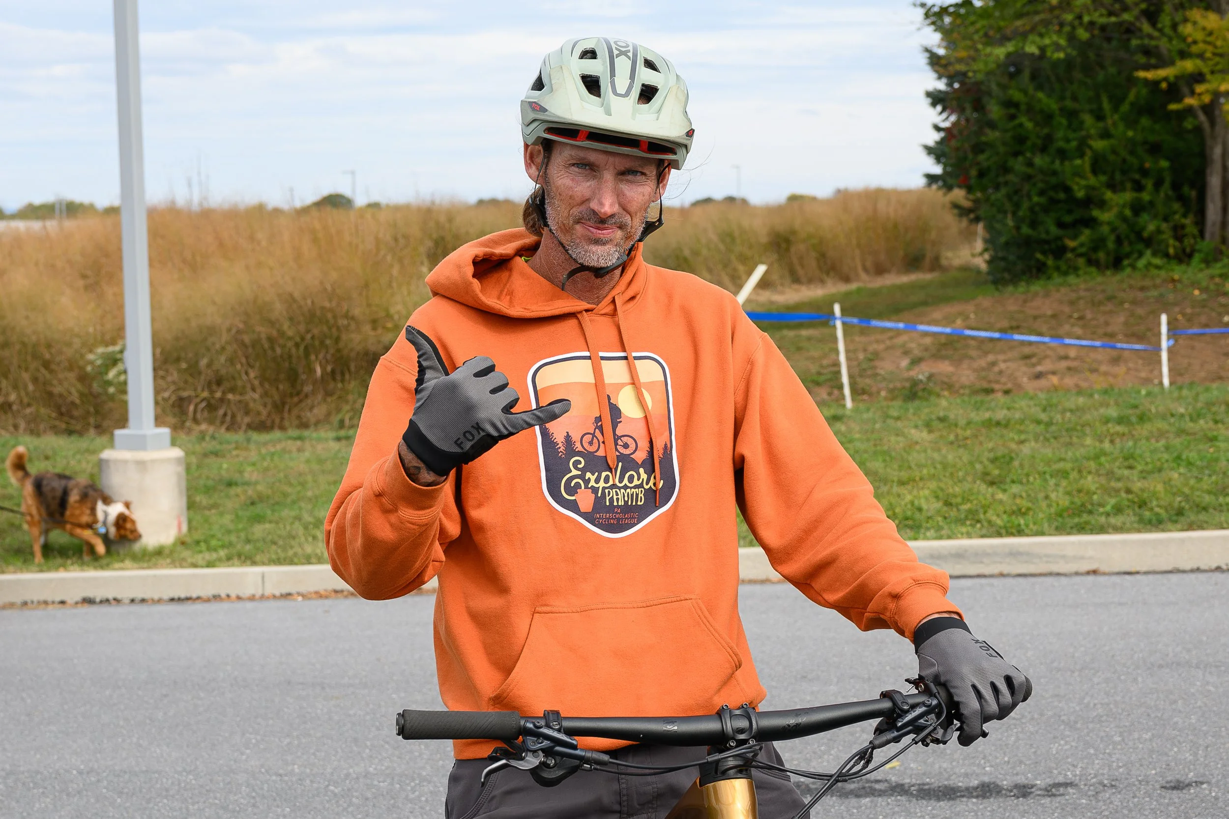 A man in an orange hoodie and a helmet standing with a bicycle outdoors, making a 'call me' gesture with his hand, with grassy fields, trees, and a dog in the background.