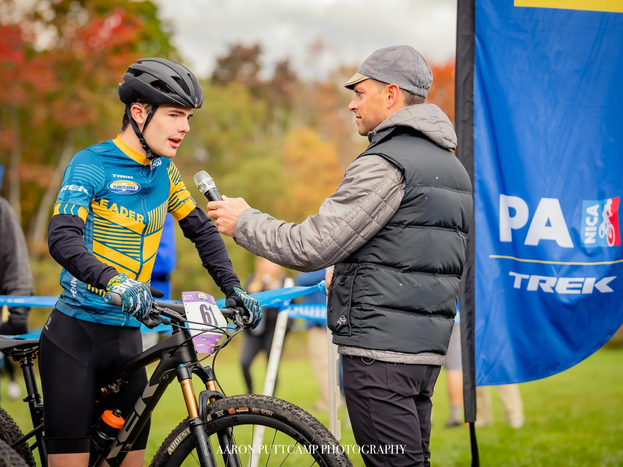 A young male mountain biker in a blue and yellow jersey being interviewed with a microphone by an older man in a gray cap and black puffy vest at an outdoor event. The cyclist is holding a mountain bike and the background features autumn trees and a blue banner.