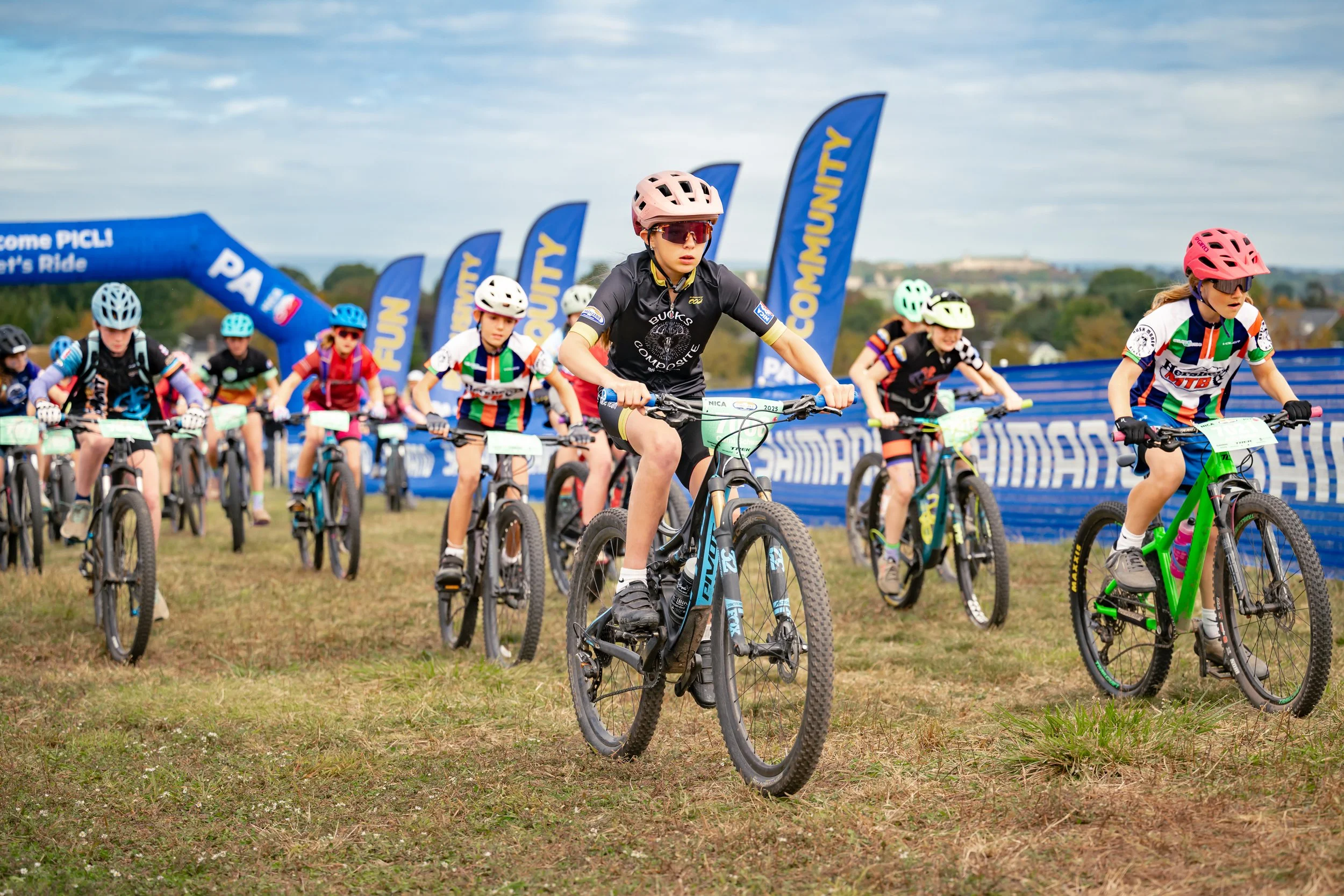 Children participating in a mountain biking race on a grassy field, wearing helmets and colorful jerseys, with blue banners and flags in the background.