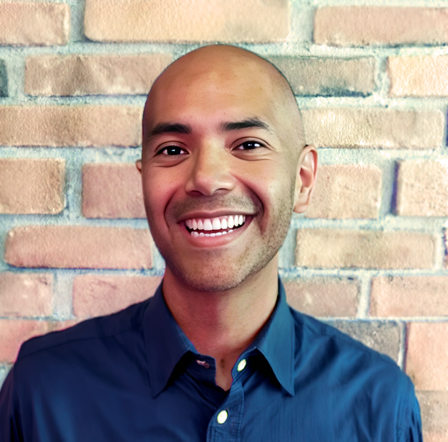 Smiling man with a shaved head wearing a navy blue collared shirt standing in front of a brick wall.