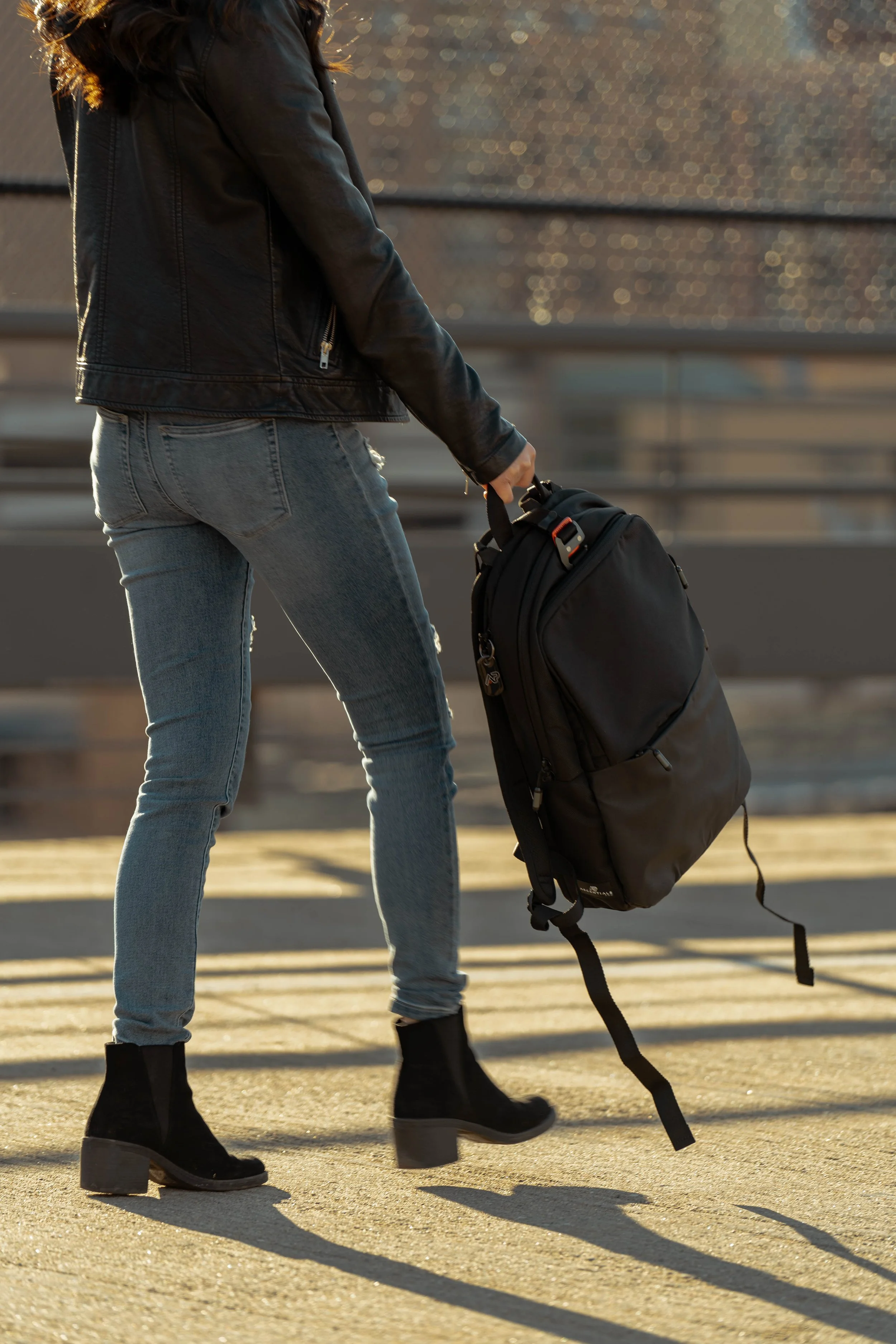 A person walking outdoors on a sunny day, carrying a black backpack, wearing black ankle boots, light blue jeans, and a black leather jacket.
