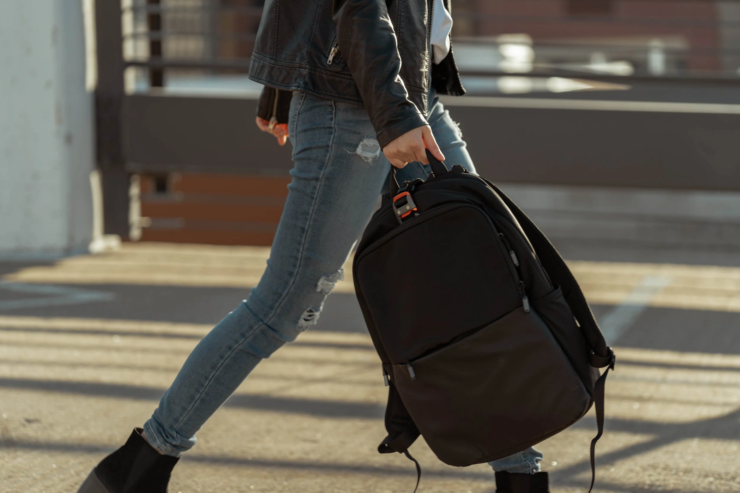 Person walking outdoors carrying a black backpack, wearing ripped jeans and a black leather jacket.