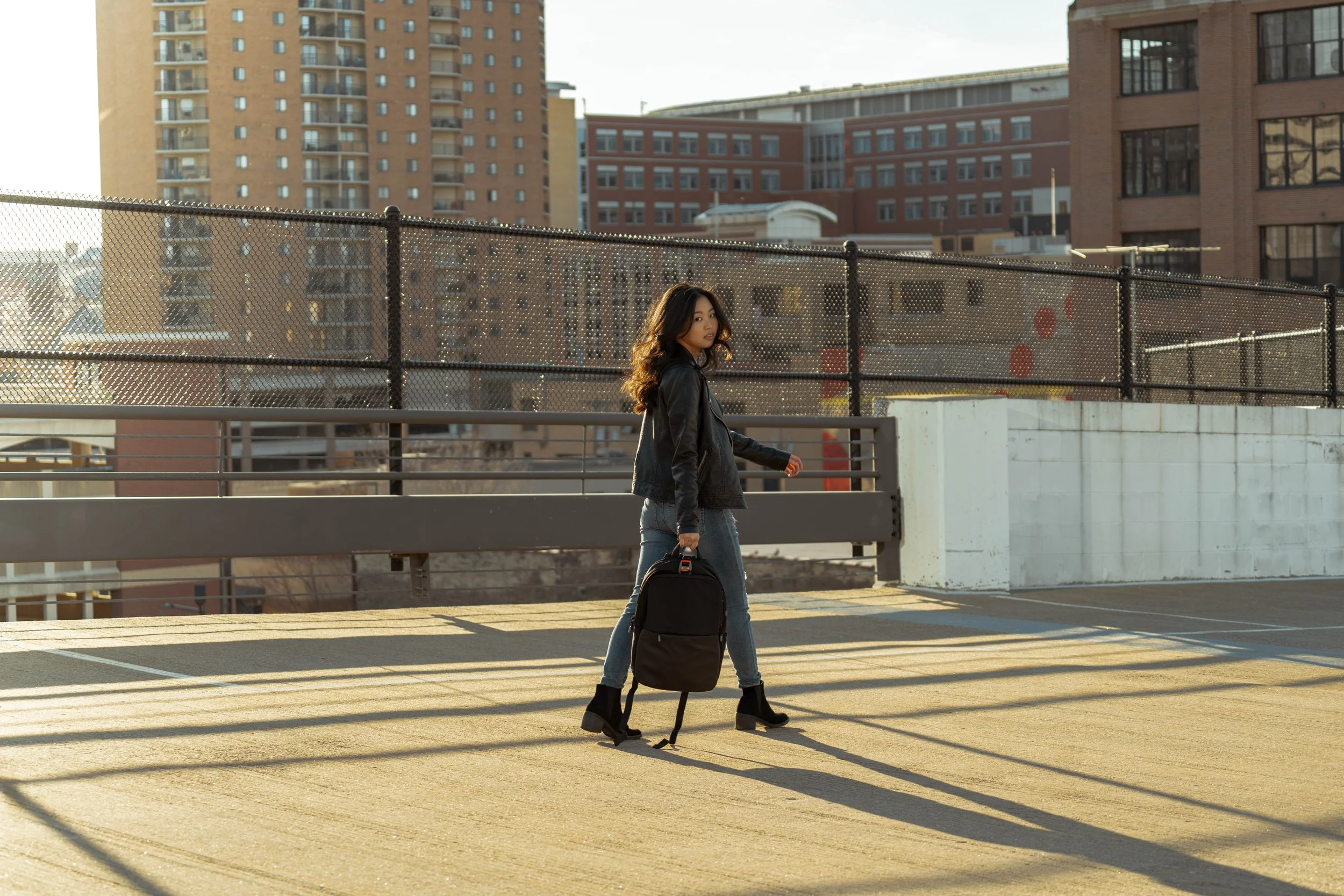 A young woman with shoulder-length hair, wearing a black leather jacket, jeans, and black ankle boots, walks across an outdoor parking garage carrying a black backpack in one hand. The setting is urban with tall buildings and a chain-link fence in th