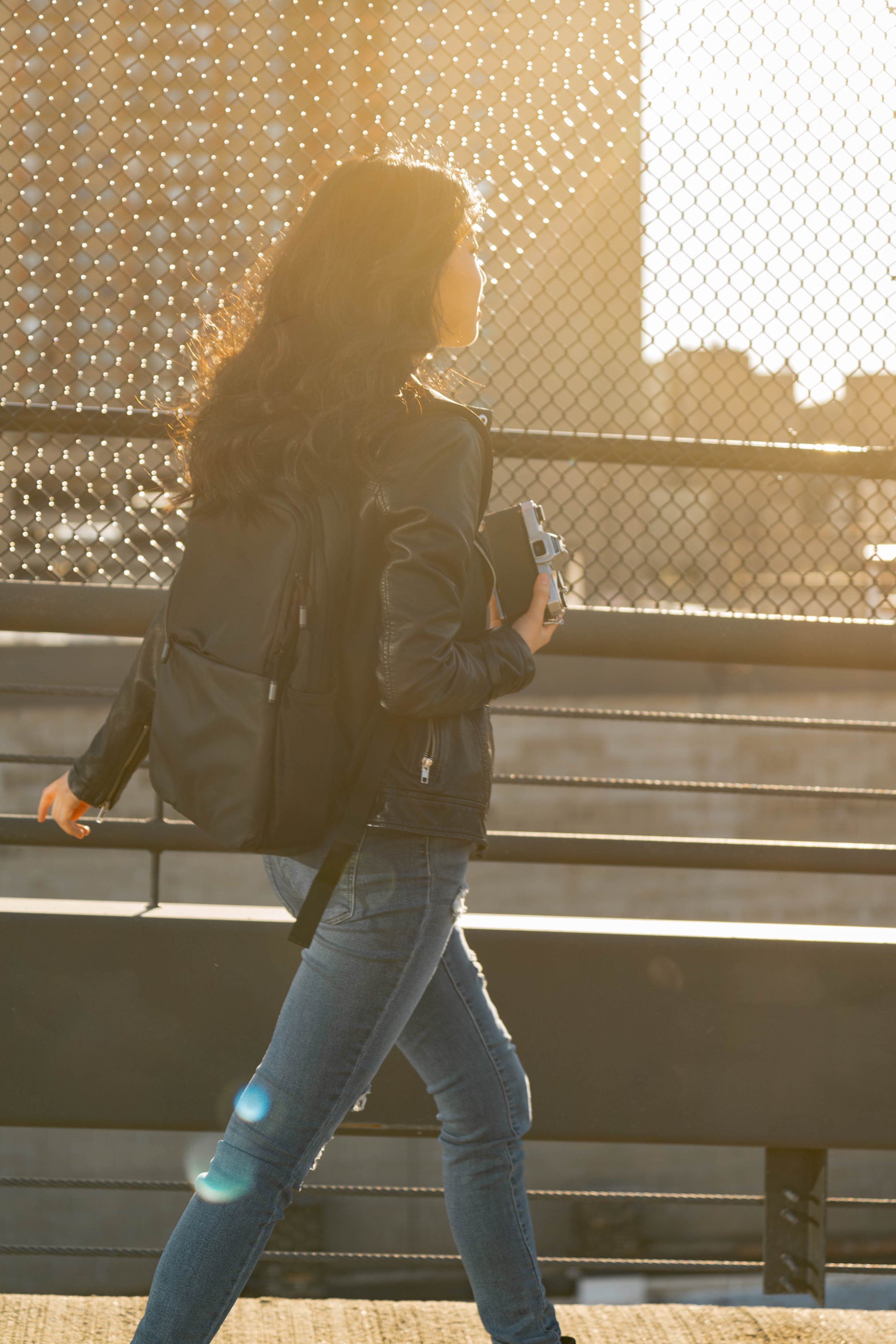 A woman with wavy hair wearing a black jacket and jeans walking on a bridge holding a small camera, with sunlight creating a warm glow.