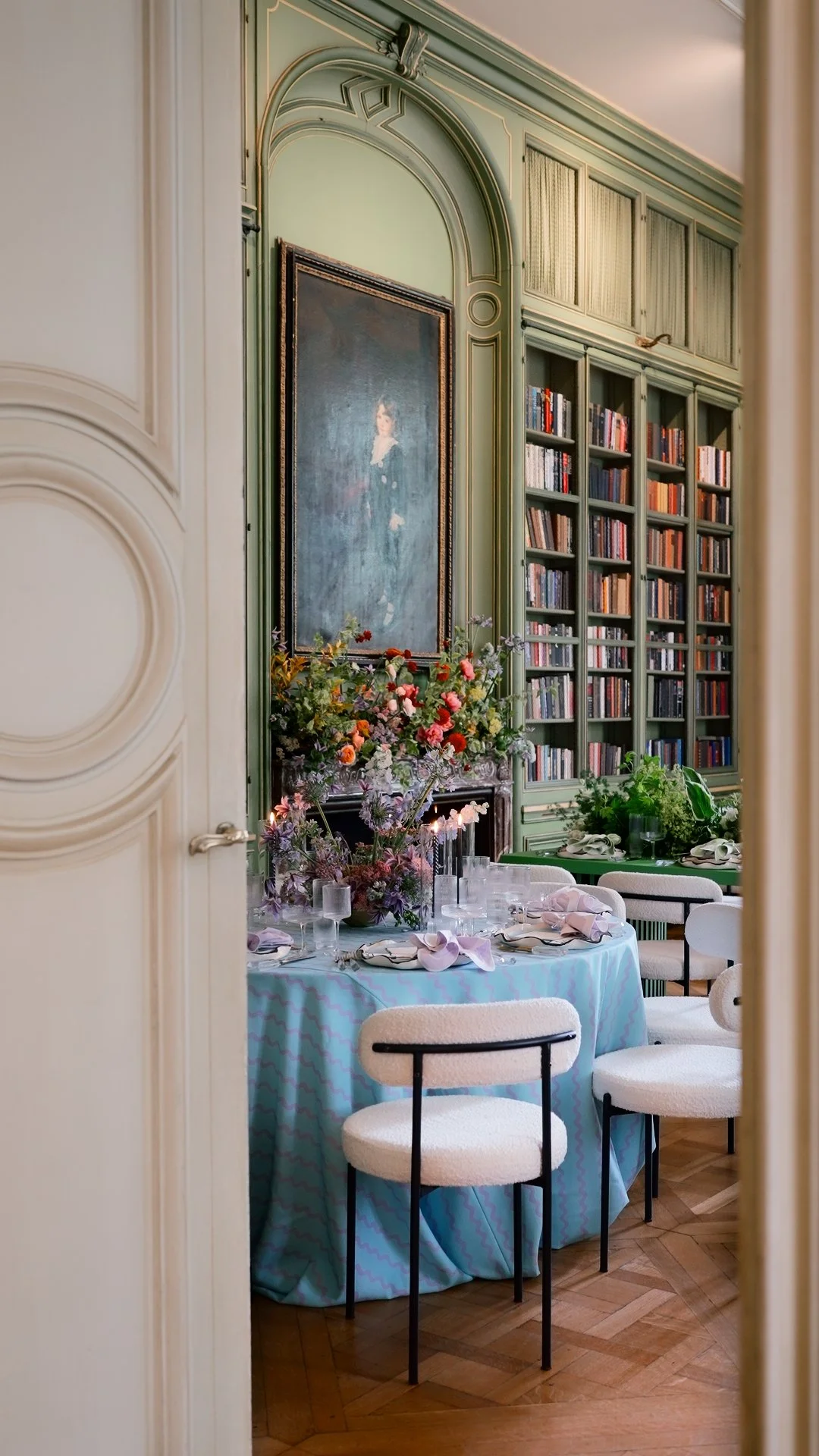 Elegant dining room with a round table covered in a pastel tablecloth, set with pink napkins, glassware, and floral centerpieces. The room features ornate molding, a large portrait, and a bookshelf filled with books, with chairs around the table and lush greenery in the background.