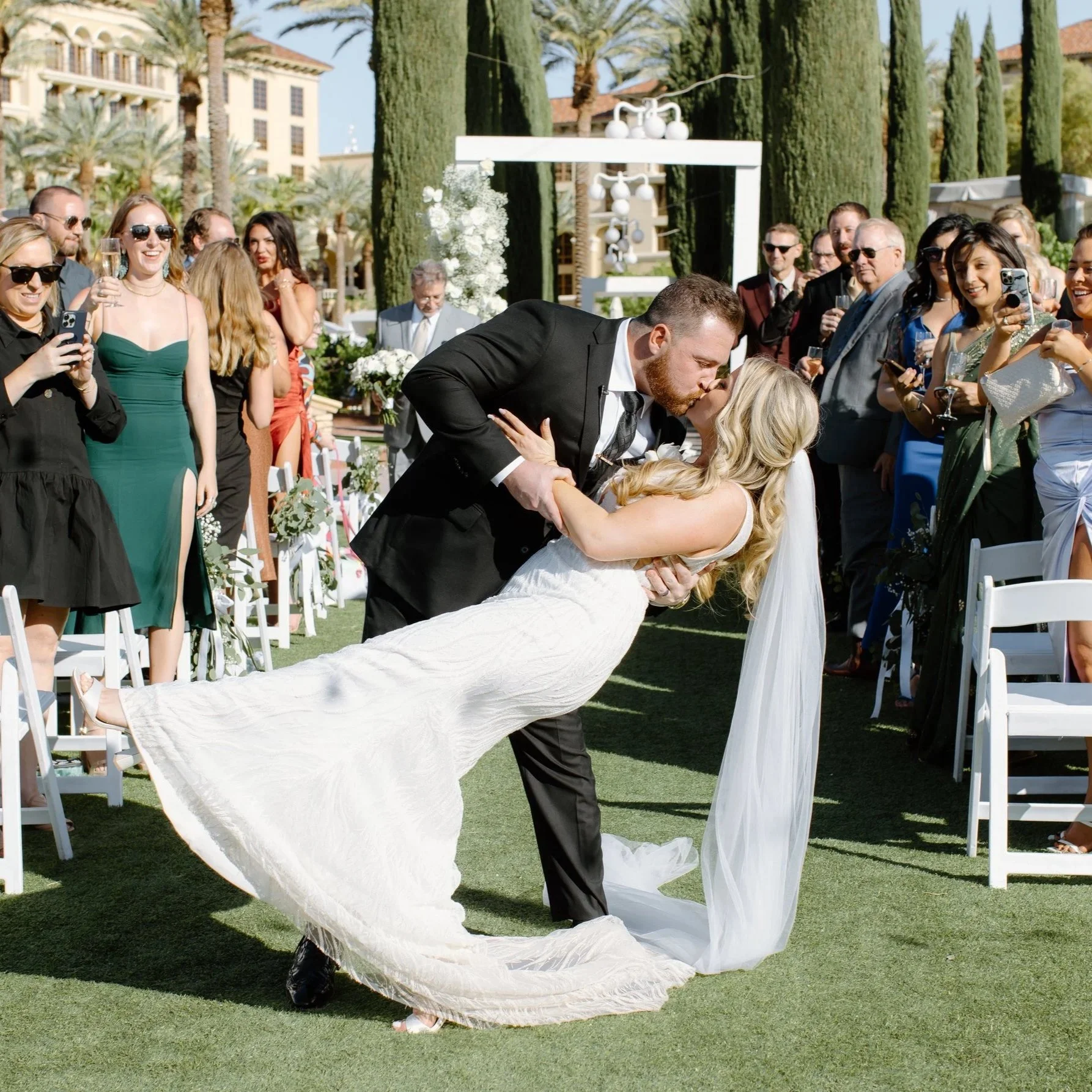 A bride and groom exchange a kiss during their outdoor wedding ceremony, surrounded by guests on a sunny day with palm trees and buildings in the background.