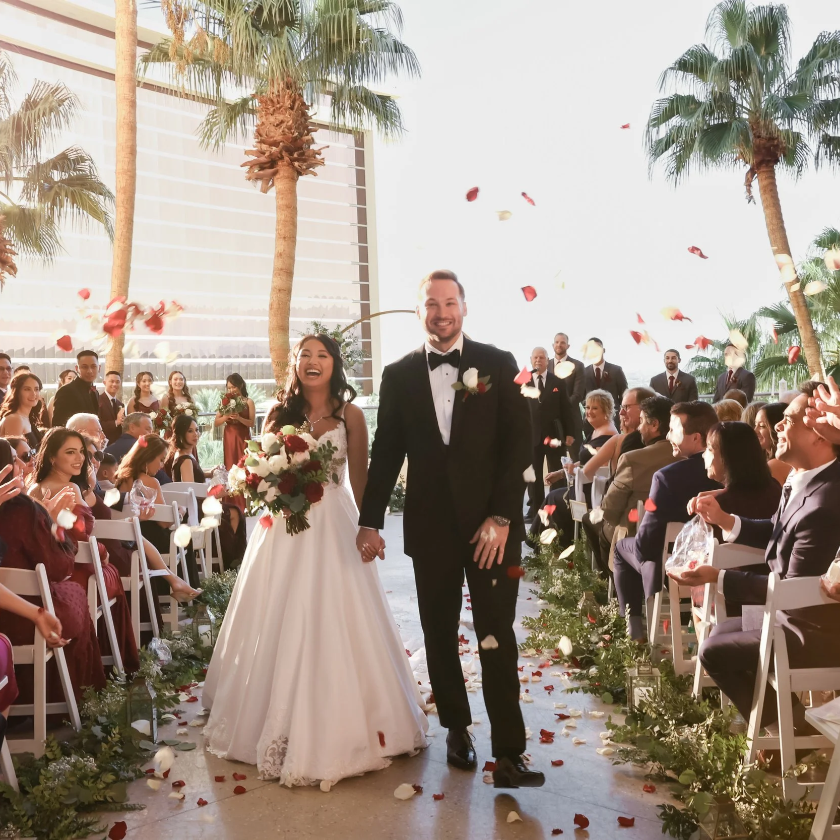 A bride and groom walking down an aisle at a wedding ceremony outdoors, surrounded by guests who are throwing flower petals. Tall palm trees and a modern building are visible in the background.