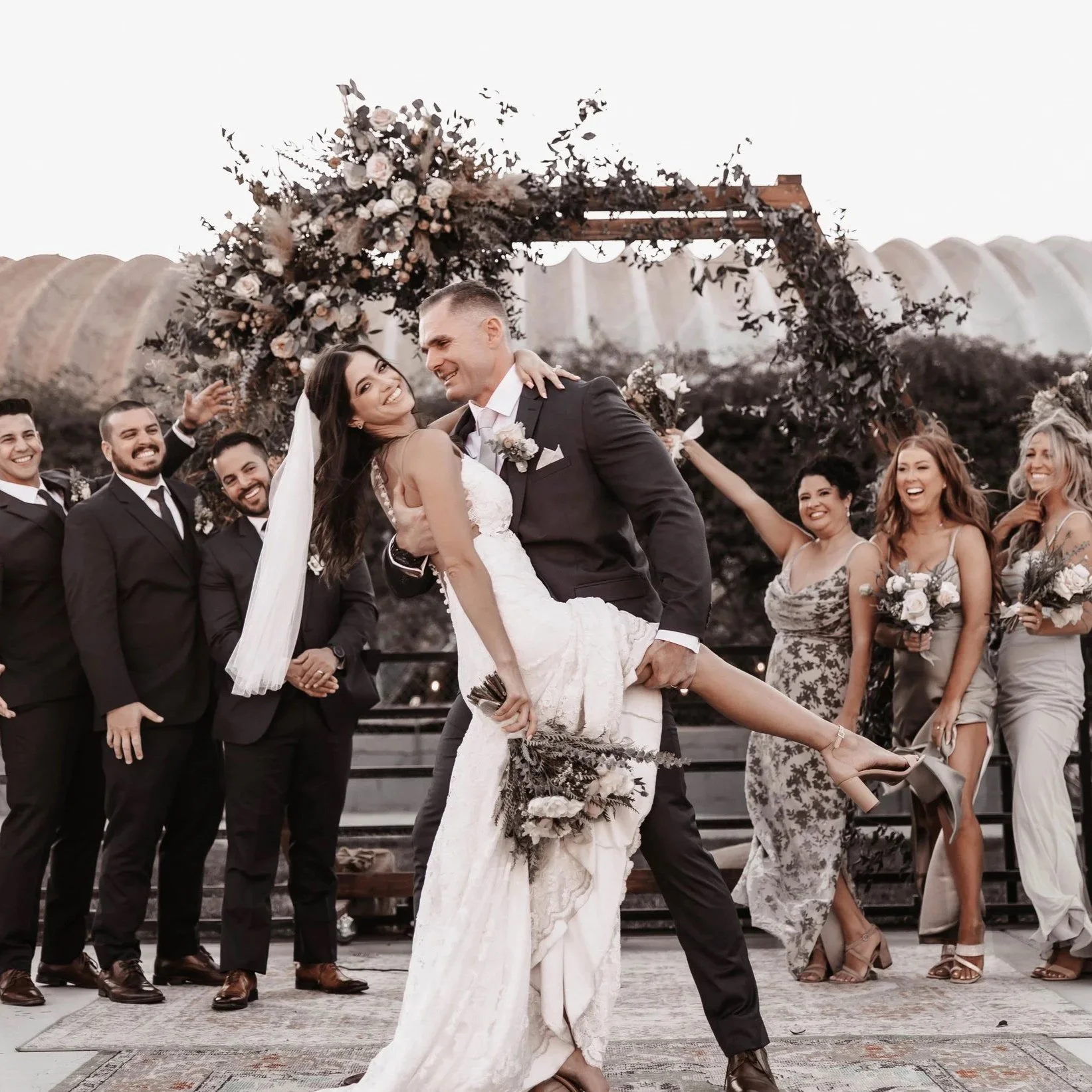 A wedding celebration with a bride and groom in the center, dancing and smiling, surrounded by friends and family outdoors with an arch decorated with flowers behind them.