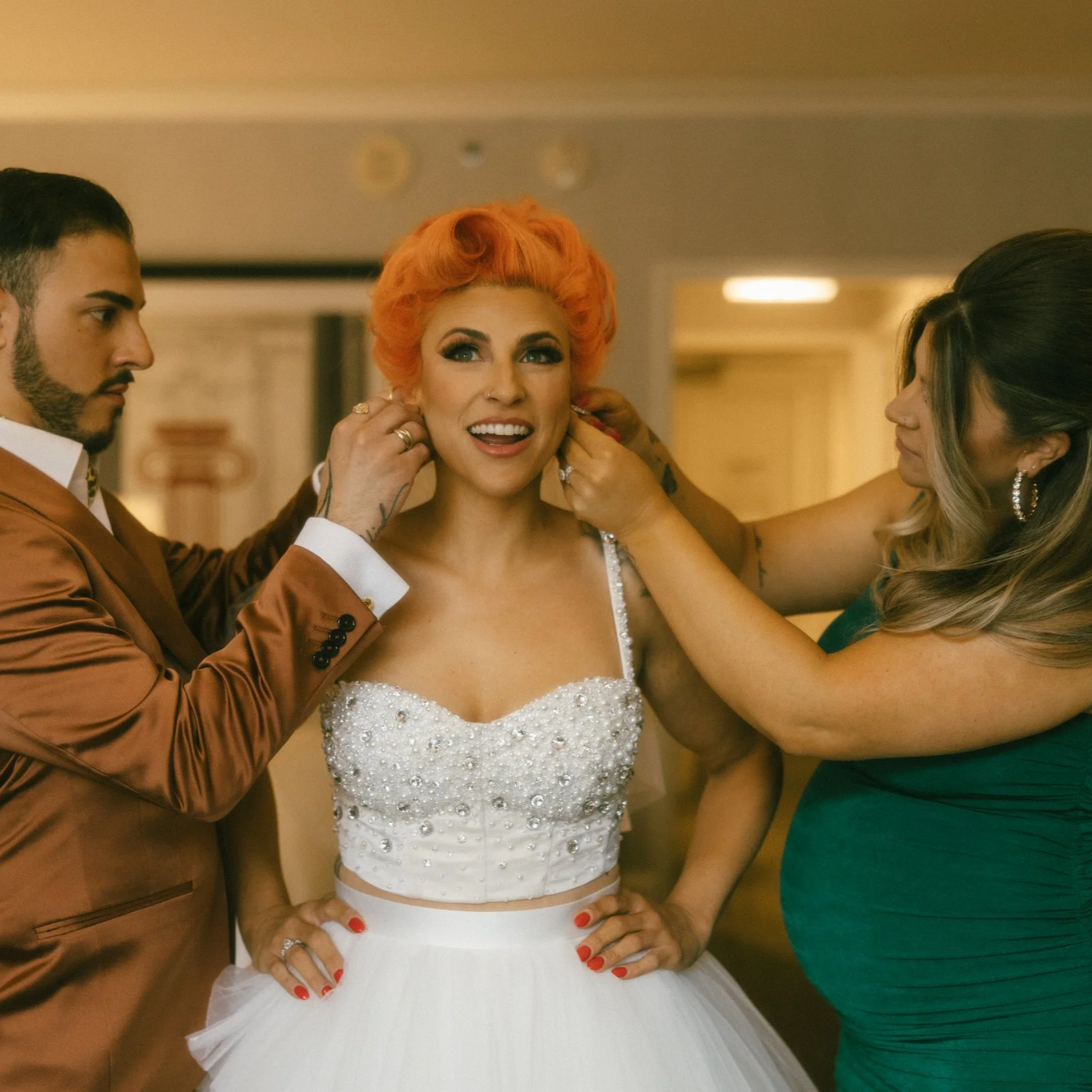 A woman in a white wedding dress is being assisted with earrings by two women, one on each side, in a warmly lit room.