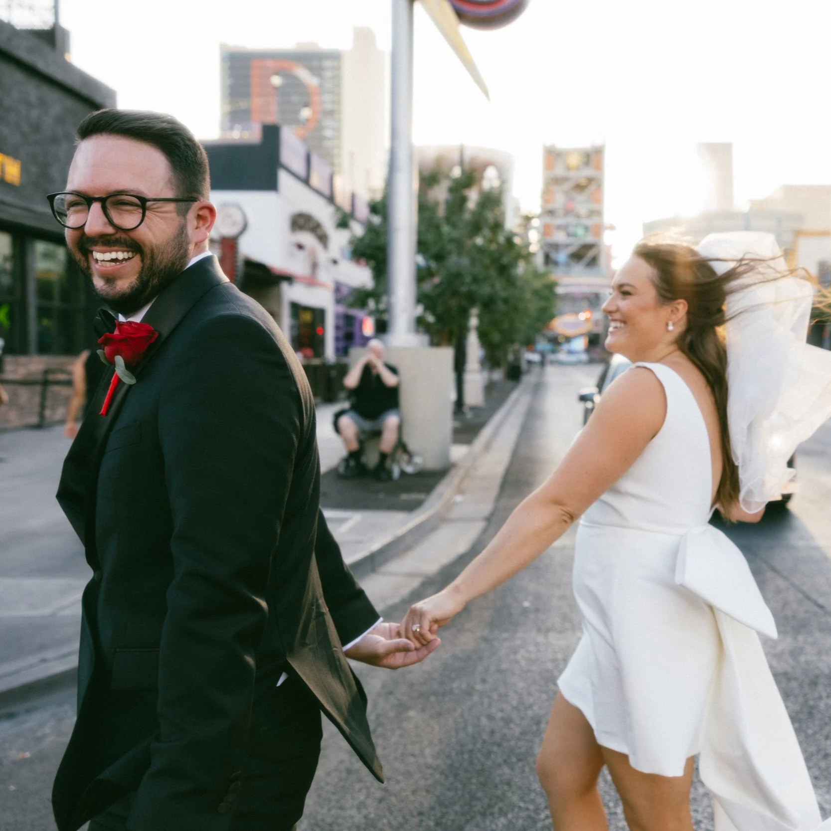 A happy couple holding hands and walking outdoors in an urban area during the daytime. The man is wearing a black suit with a red boutonniere, glasses, and has a beard. The woman is wearing a white dress with a large bow in her hair. They are smiling