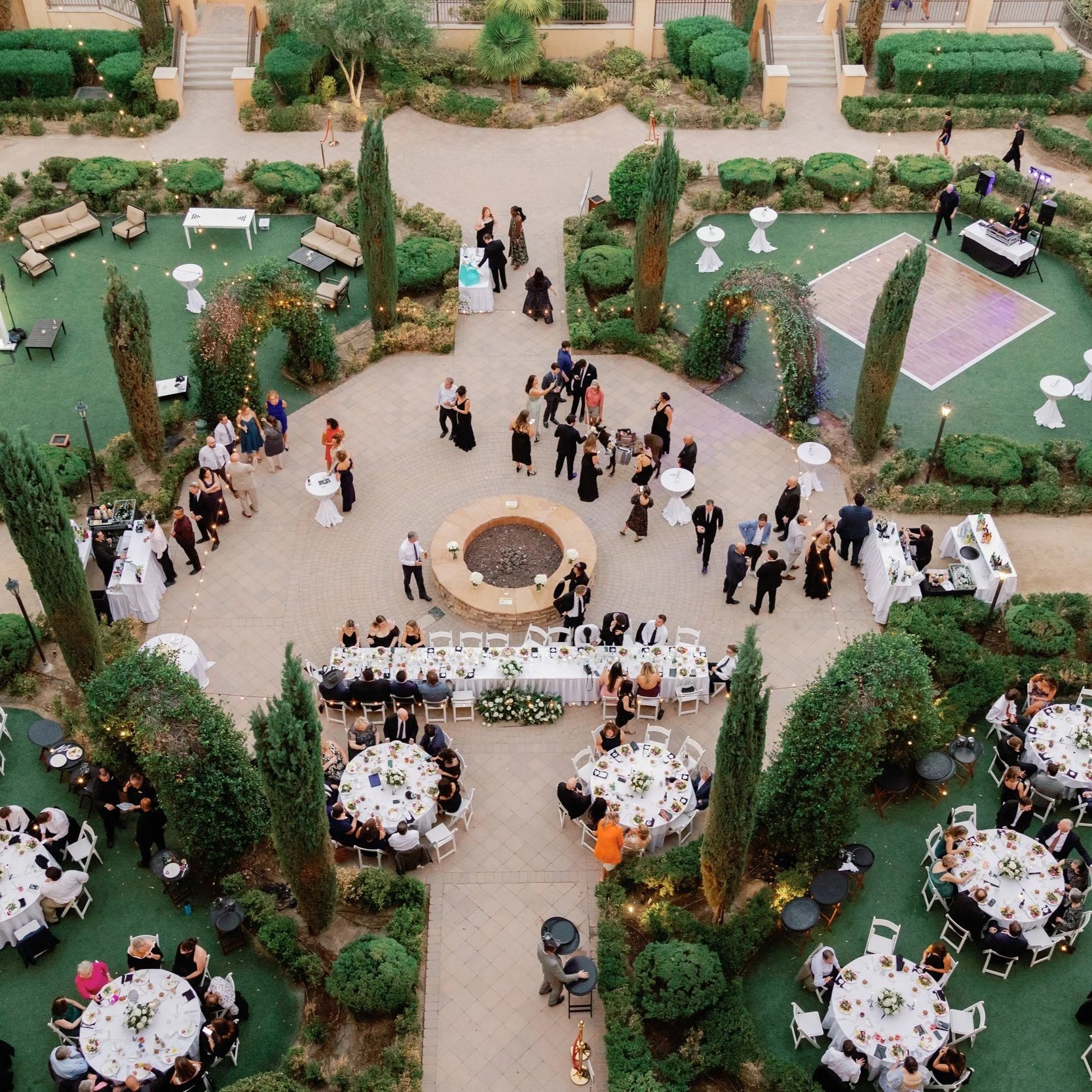 An aerial view of an elegant outdoor wedding reception set in a lush garden with numerous round dining tables decorated with flowers, people mingling and dancing, and a DJ setup with speakers on the right side.
