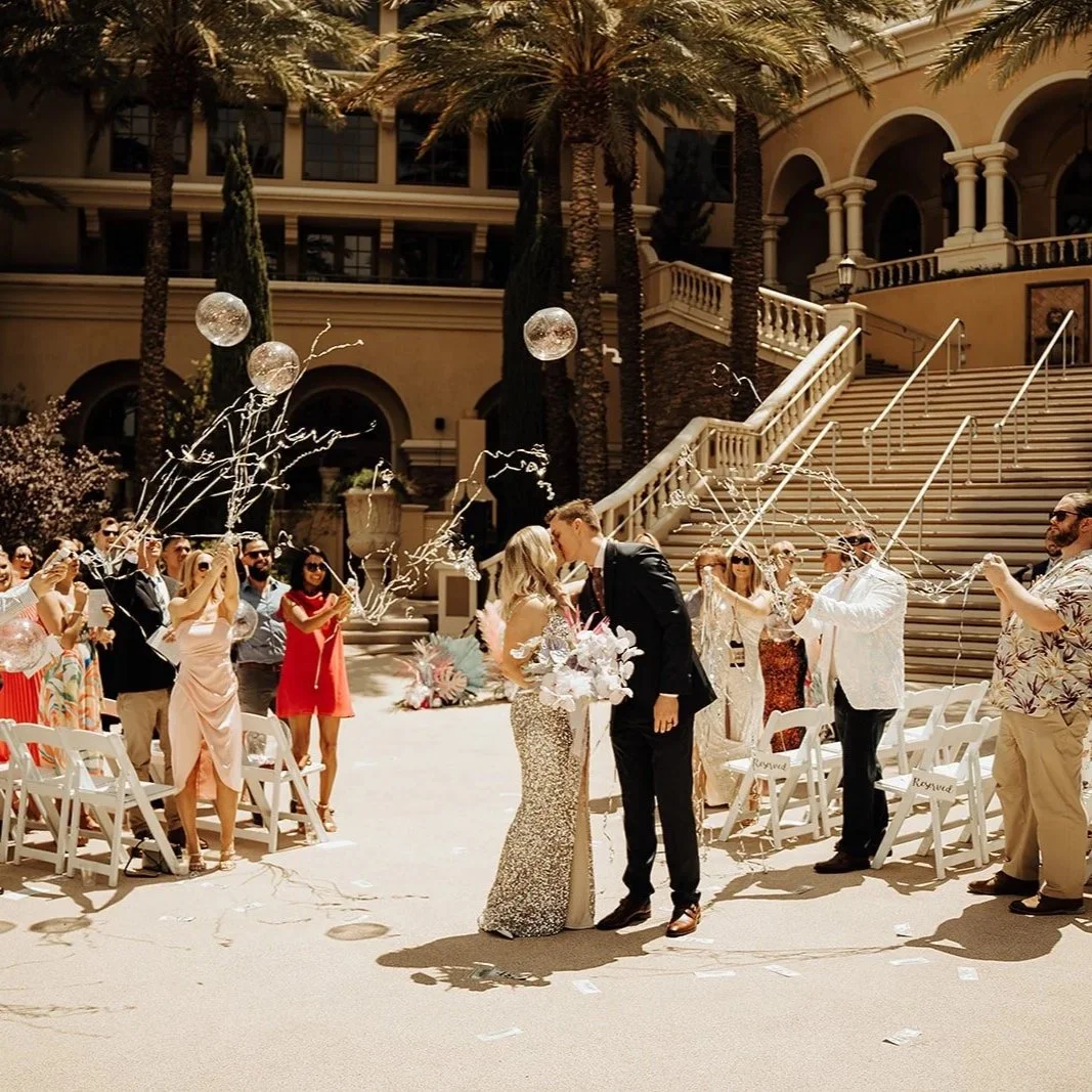 A wedding celebration outdoors with a bride and groom kissing in the center, guests throwing balloons, confetti, and streamers, on a sunny day with a grand staircase and palm trees in the background.