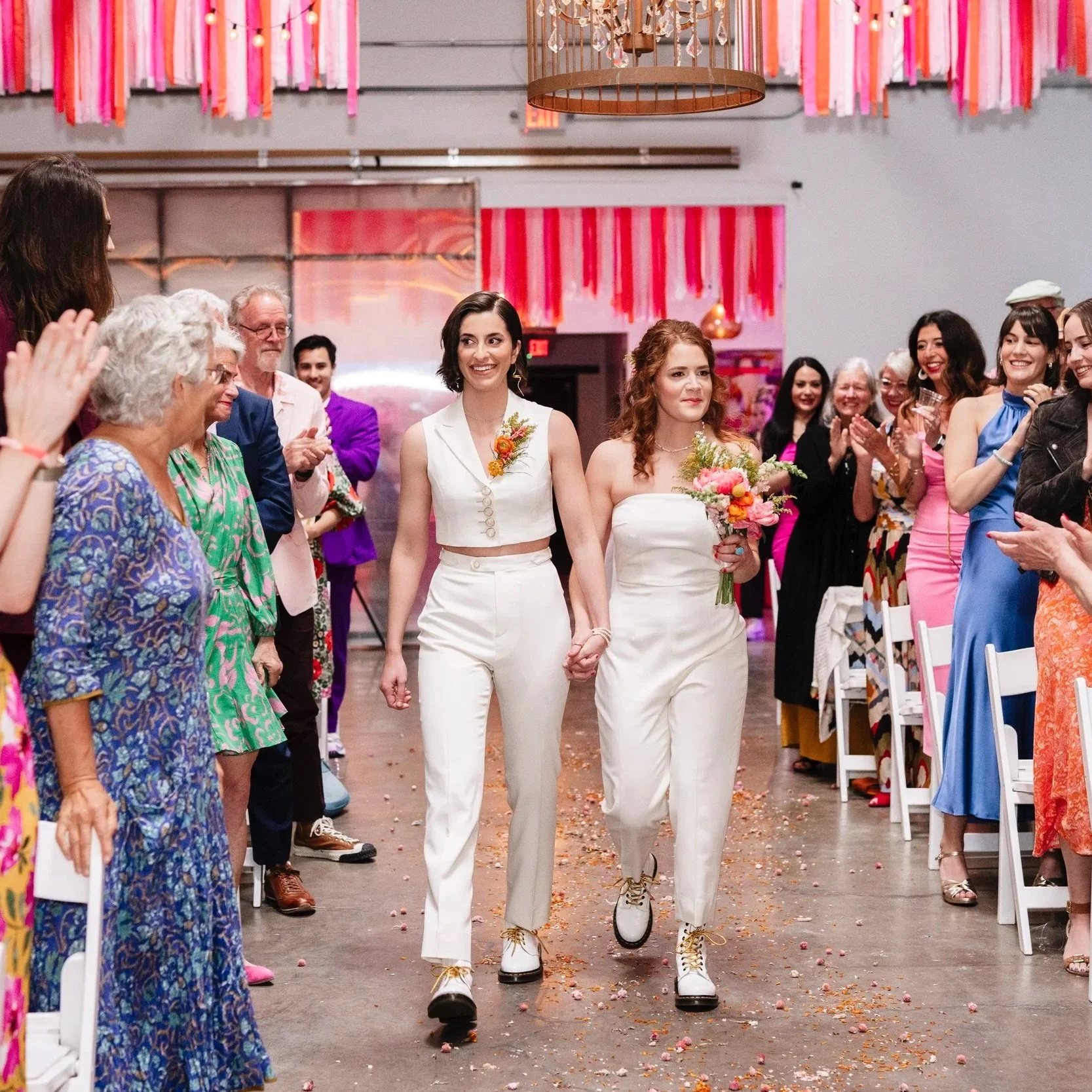 Two women, one in a white pantsuit with flowers and one in a white dress, walk down an aisle holding hands at a celebration, surrounded by guests and colorful decorations.