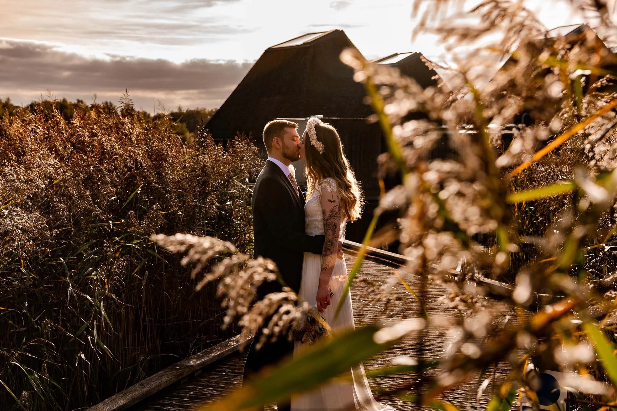 A bride and groom sharing a kiss on a wooden walkway surrounded by tall grasses at sunset in the rural setting of Brockholes Nature Reserve.